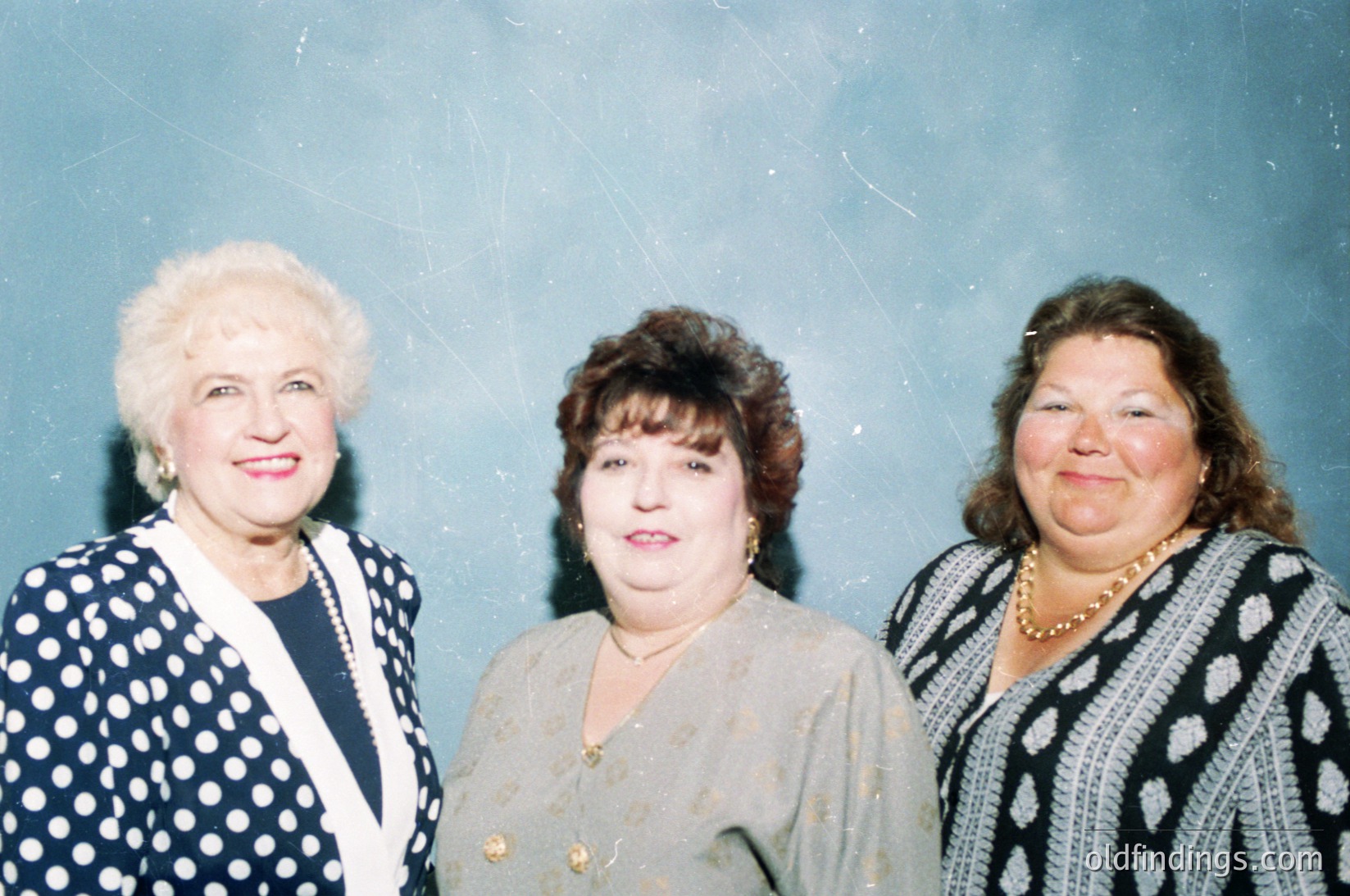 Three women in formal attire pose indoors, likely mid-1990s to early 2000s. Left: white-blonde hair, dark polka-dot blazer with pearl earrings. Center: dark bob haircut, beige blouse with gold brooch. Right: wavy brown hair, striped sweater with pearl necklace. Background suggests a formal event or office setting.
