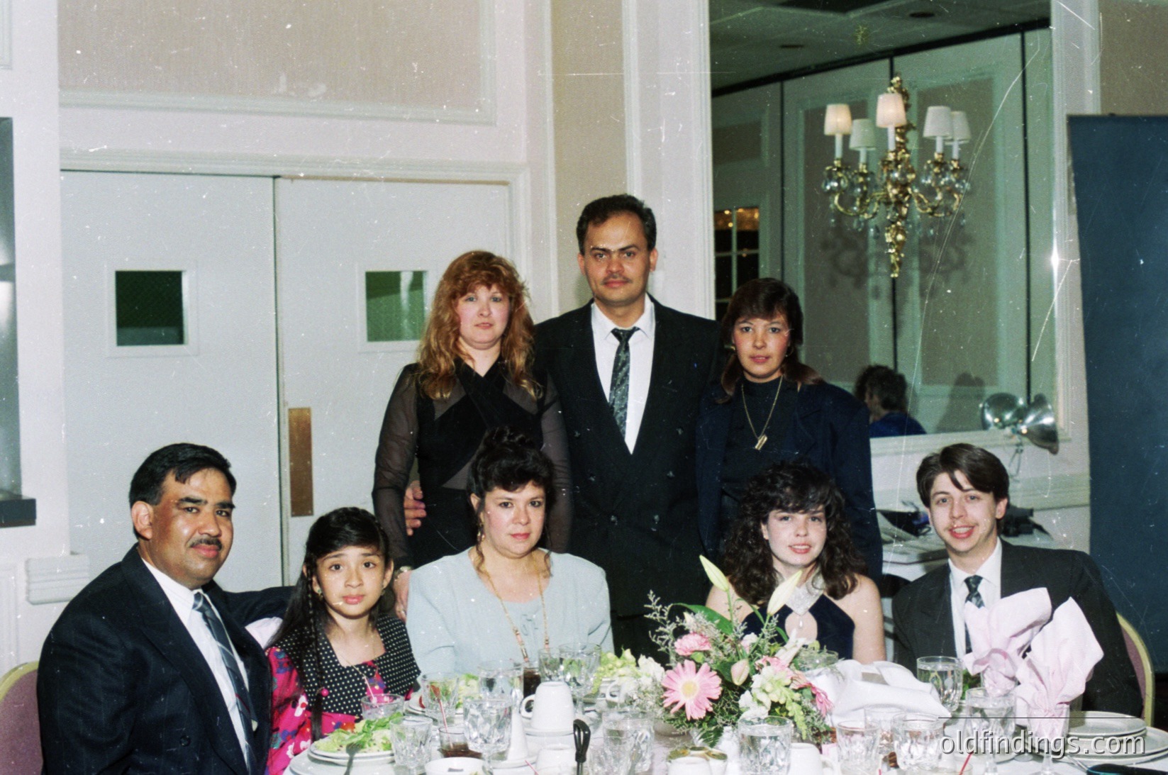 Family portrait at a formal banquet hall, likely 1980s–1990s. Seven individuals pose indoors with a chandelier and mirrored wall backdrop. Men wear suits, women in formal dresses; floral centerpieces adorn the table.
