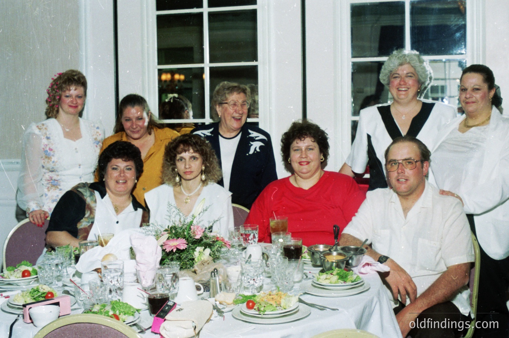 Family gathering at a formal indoor banquet, likely 1990s. Eight adults pose around a long table set with crystal glasses, floral centerpieces, and place settings. Women wear blouses with floral/patterned designs; men in dress shirts. Soft lighting and large windows suggest an elegant venue.