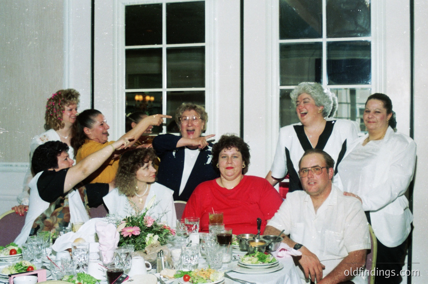 Elegant 1990s indoor celebration with nine guests seated at a long banquet table adorned with floral centerpieces, wine glasses, and plates. Formal attire—women in dresses, men in suits—suggests a wedding or anniversary. Bright lighting and large windows indicate a spacious venue. Candid smiles and gestures convey joy.