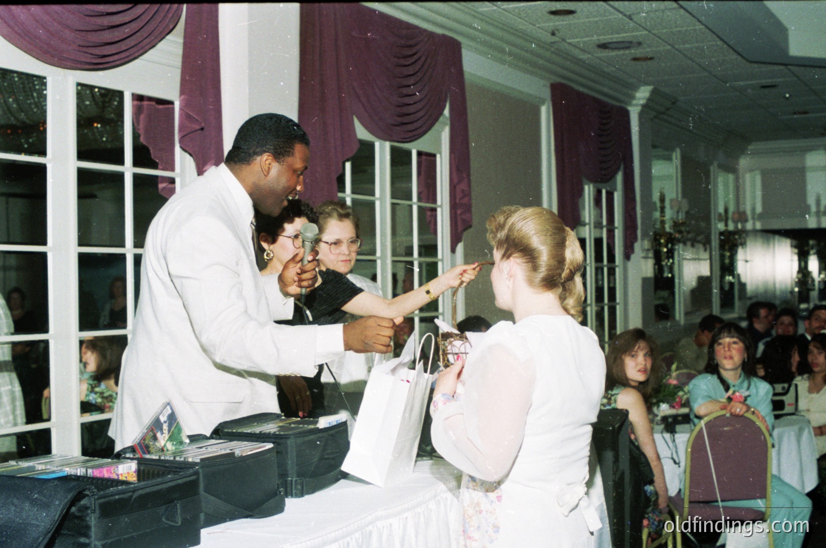 Vintage banquet hall scene featuring a male waiter in white uniform handing a bridal bouquet to a bride in 1970s attire. Purple drapes and floral table settings suggest a formal event. Crowd in background includes guests in semi-formal wear.