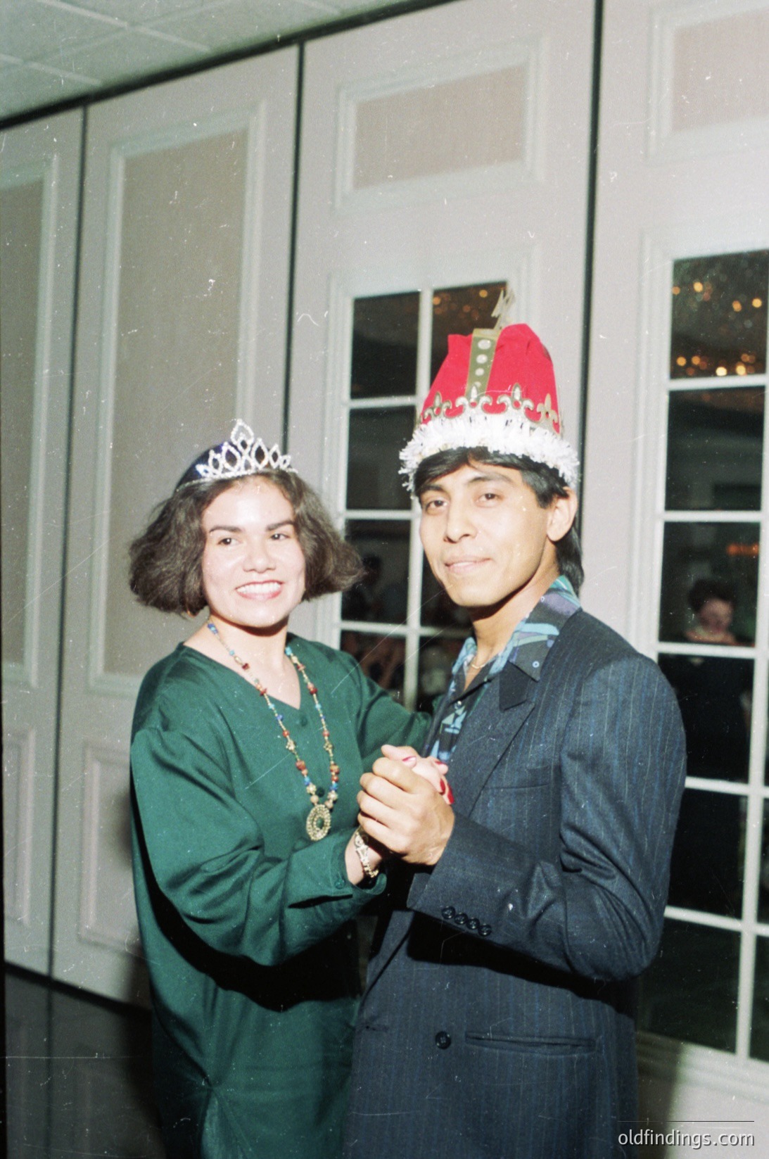 Two individuals pose indoors, likely at a festive event in the 1970s–1980s. The woman wears a tiara and green dress with a beaded necklace, while the man dons a Santa hat with gold trim and a dark suit. Reflections in windows suggest a formal venue, possibly a holiday party or themed gathering.