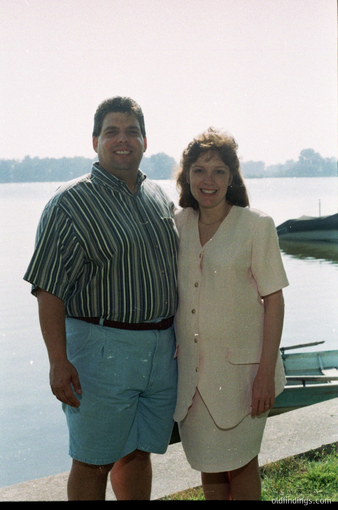 Couple posing by a serene lakefront, 1980s-1990s. Man in striped button-down shirt and rolled cuff shorts; woman in sleeveless beige blouse with button details. Boat docked in background, lush greenery.
