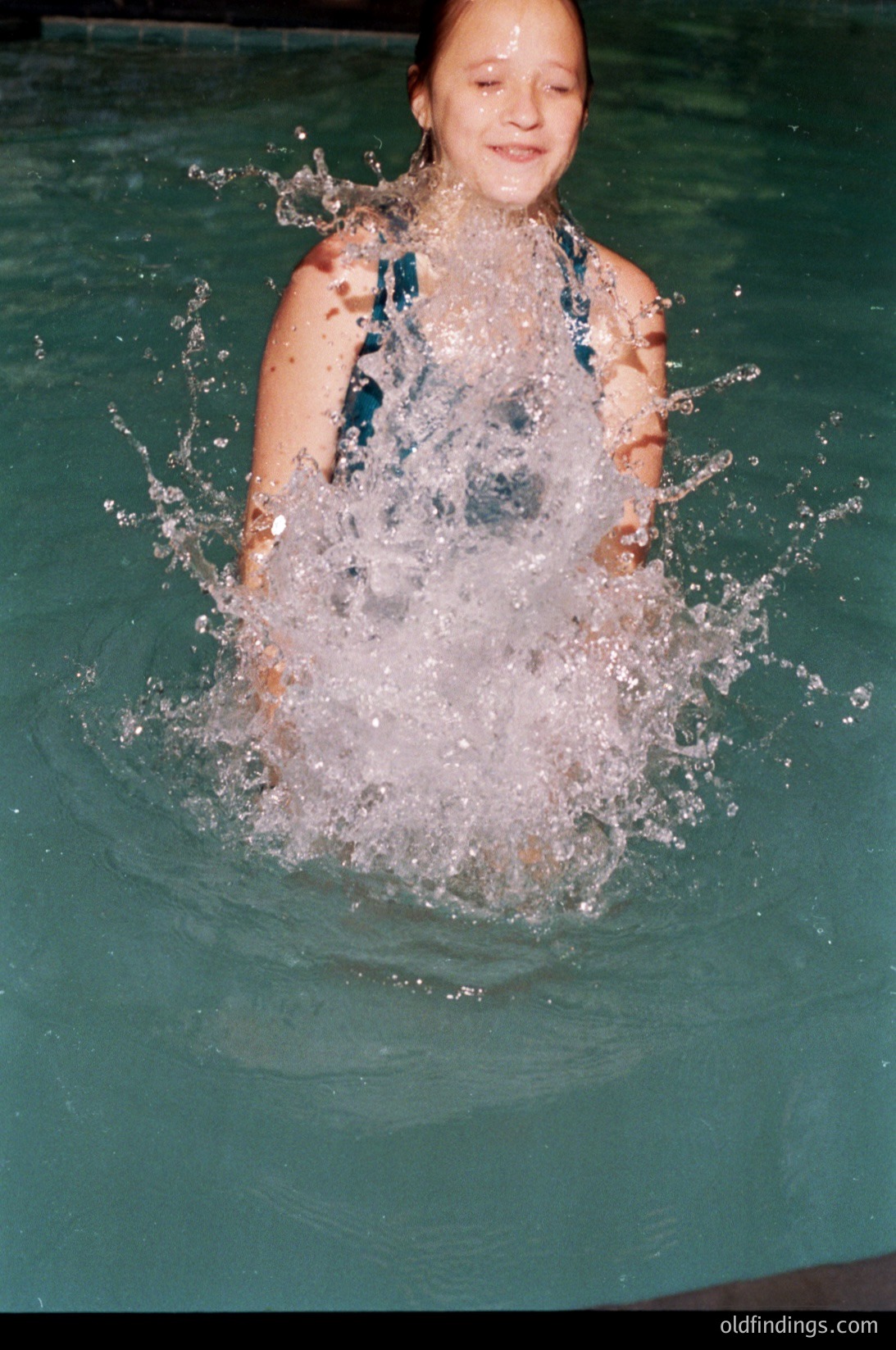 Young girl mid-dive in an indoor pool, creating a splash. Wearing a blue one-piece swimsuit with a textured pattern. Likely mid-1990s to 2000s based on swimwear style and image quality.