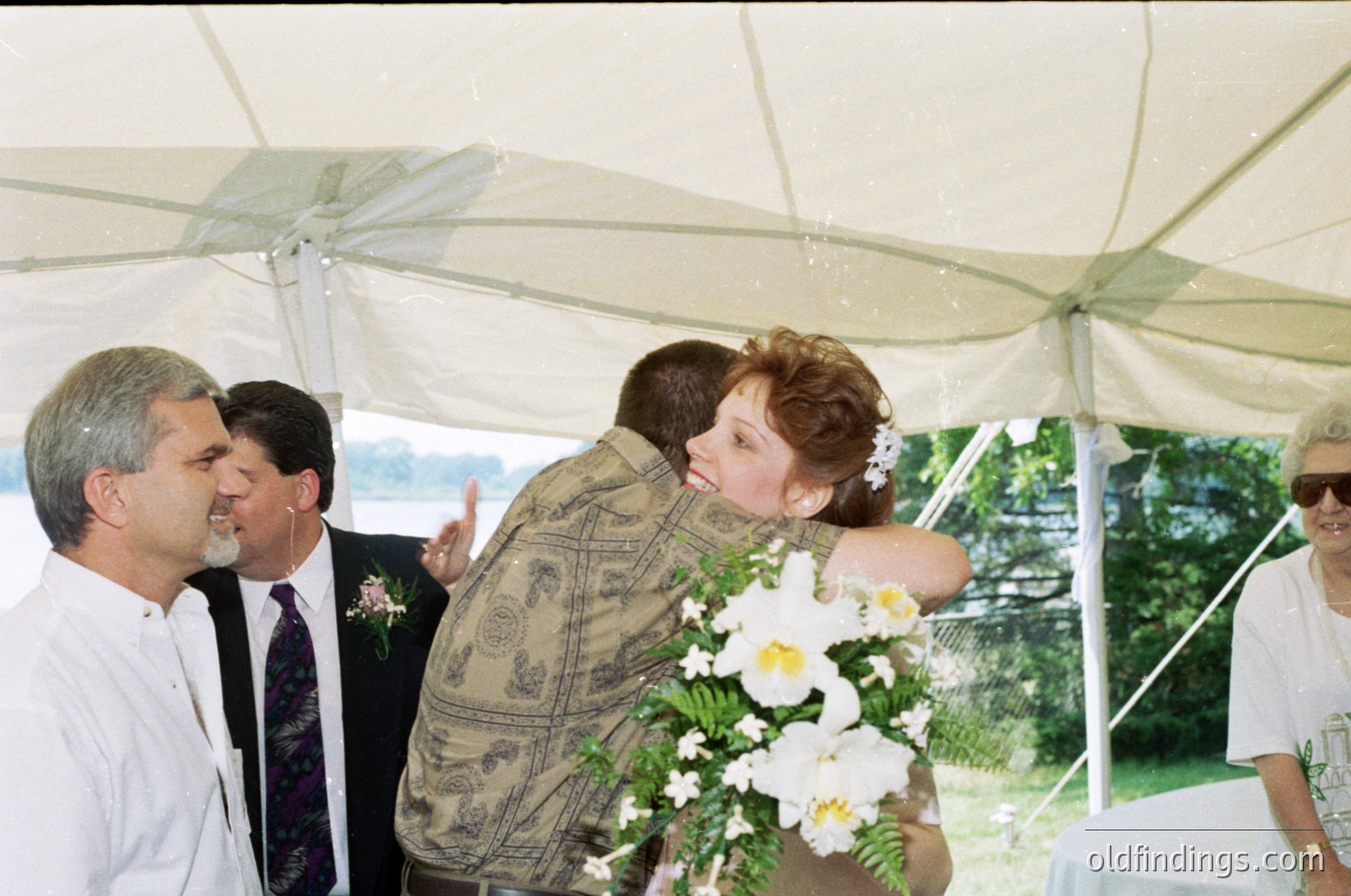 Under a white canopy tent, a bride in a floral dress and groom in a patterned shirt share an emotional moment. Surrounding them, guests in formal attire—suits and ties—witness the exchange. The bouquet features white flowers, likely peonies. Outdoor setting suggests a garden or park venue. Likely a 1990s wedding based on attire and photography style.
