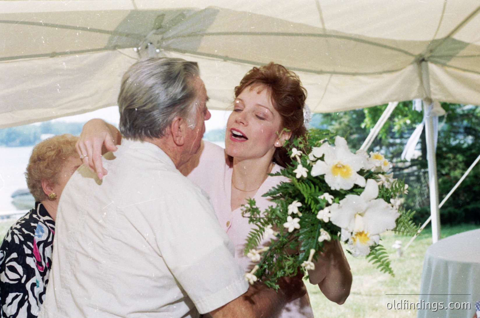 A joyous moment captured: an elderly man in a white shirt embraces a woman in a light pink dress, holding a bouquet of white flowers. The scene appears to be an outdoor celebration under a white canopy, likely a wedding or milestone event. The setting suggests a mid-20th century (1960s–1980s) Western or European location. [Elderly man hugging bride under white canopy, holding floral bouquet ]