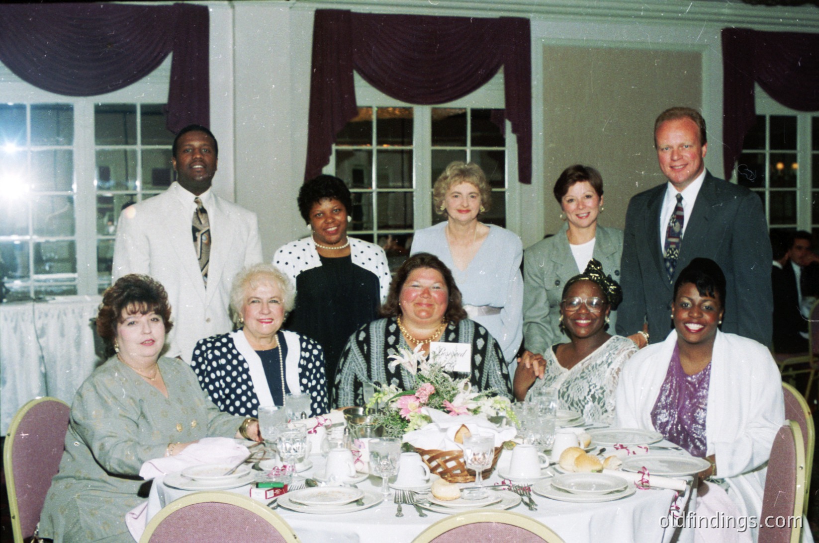 Elegant 1990s formal gathering at a banquet hall, featuring nine adults seated at a long table adorned with floral centerpieces and silverware. Attendees wear business attire—blazers, dresses, and ties—suggesting a corporate or celebratory event. Soft draping and large windows indicate a sophisticated venue.
