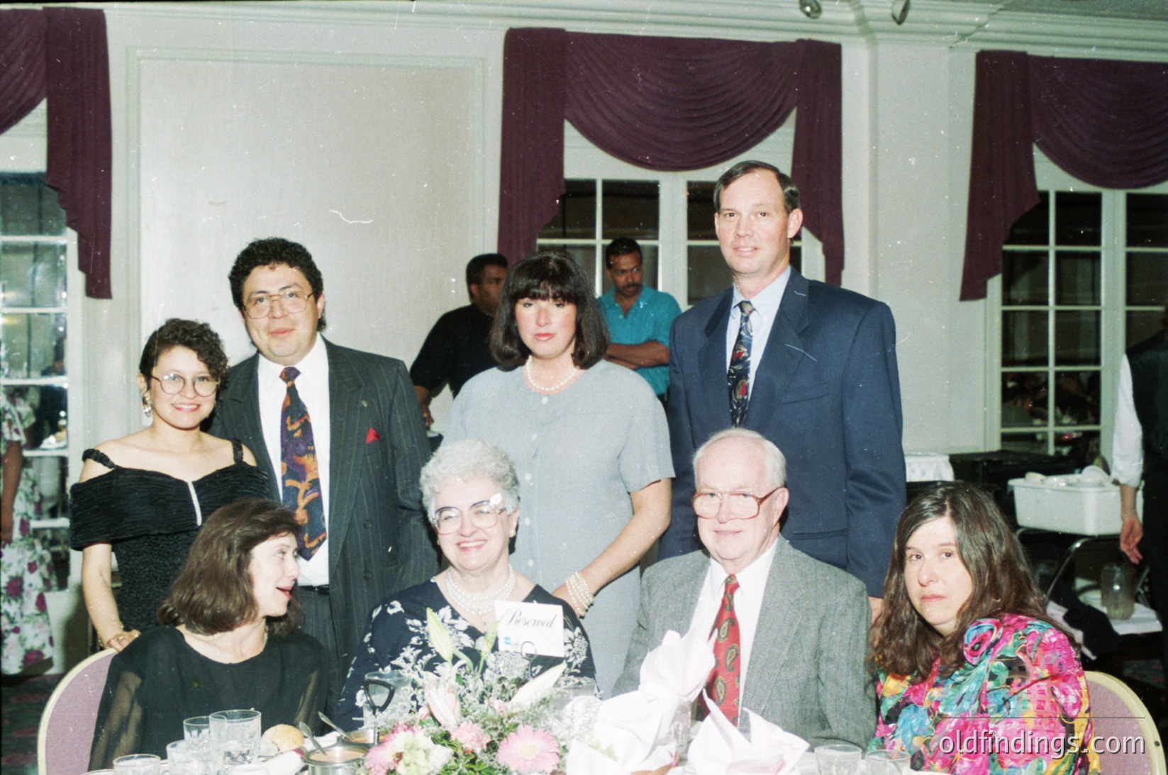 Family portrait at a formal indoor event, likely a wedding or anniversary, featuring seven adults seated and standing around a table adorned with floral arrangements. Men wear suits with ties, women in formal dresses and blouses. The setting includes draped curtains and a chandelier, suggesting a banquet hall or event venue. Style and attire suggest the 1990s.