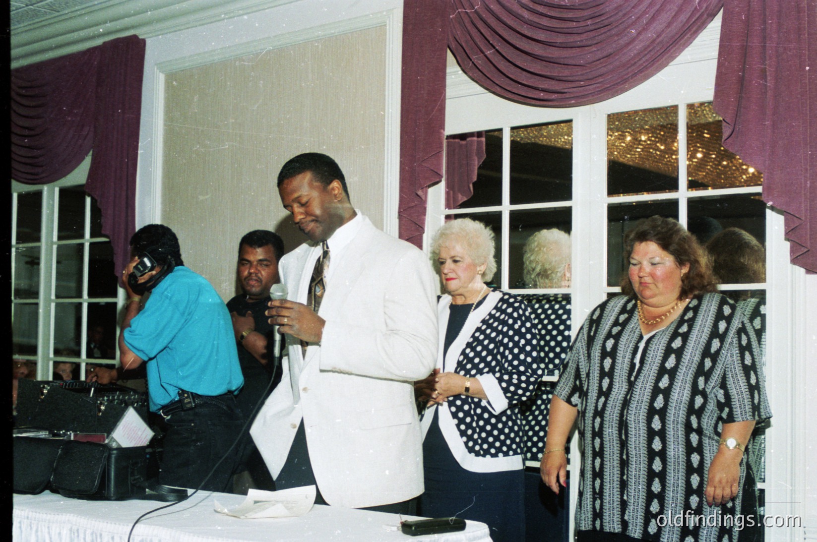 Elegant 1990s indoor event with polished wooden floors and draped maroon/purple curtains. Central figure in white suit speaking into mic, flanked by attendees in formal attire—polka-dot blazers, patterned dresses. DJ booth with equipment on left. Soft lighting enhances warm, intimate ambiance.