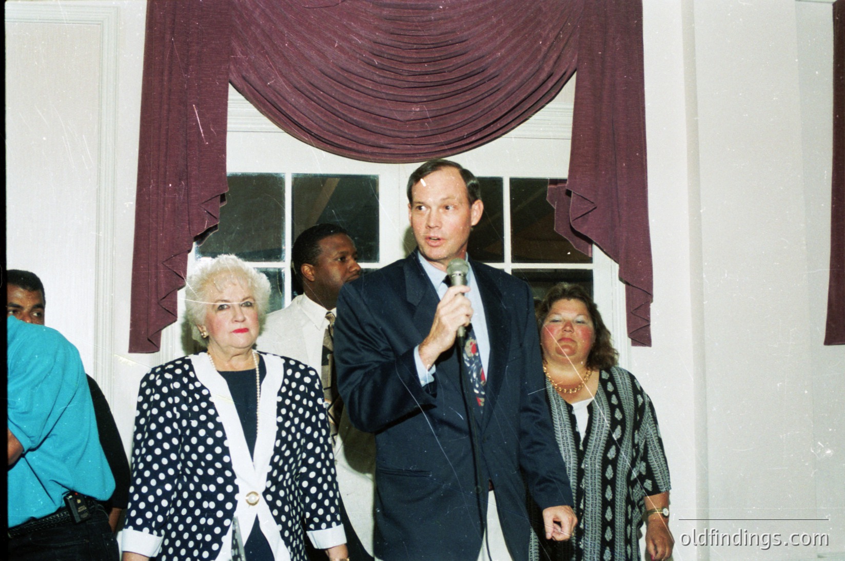 Indoor formal event featuring a man in a dark suit speaking into a microphone, flanked by three women in formal attire. The backdrop includes burgundy drapes and a white wall with a window. Clothing suggests 1980s–1990s era, likely a public or corporate gathering.