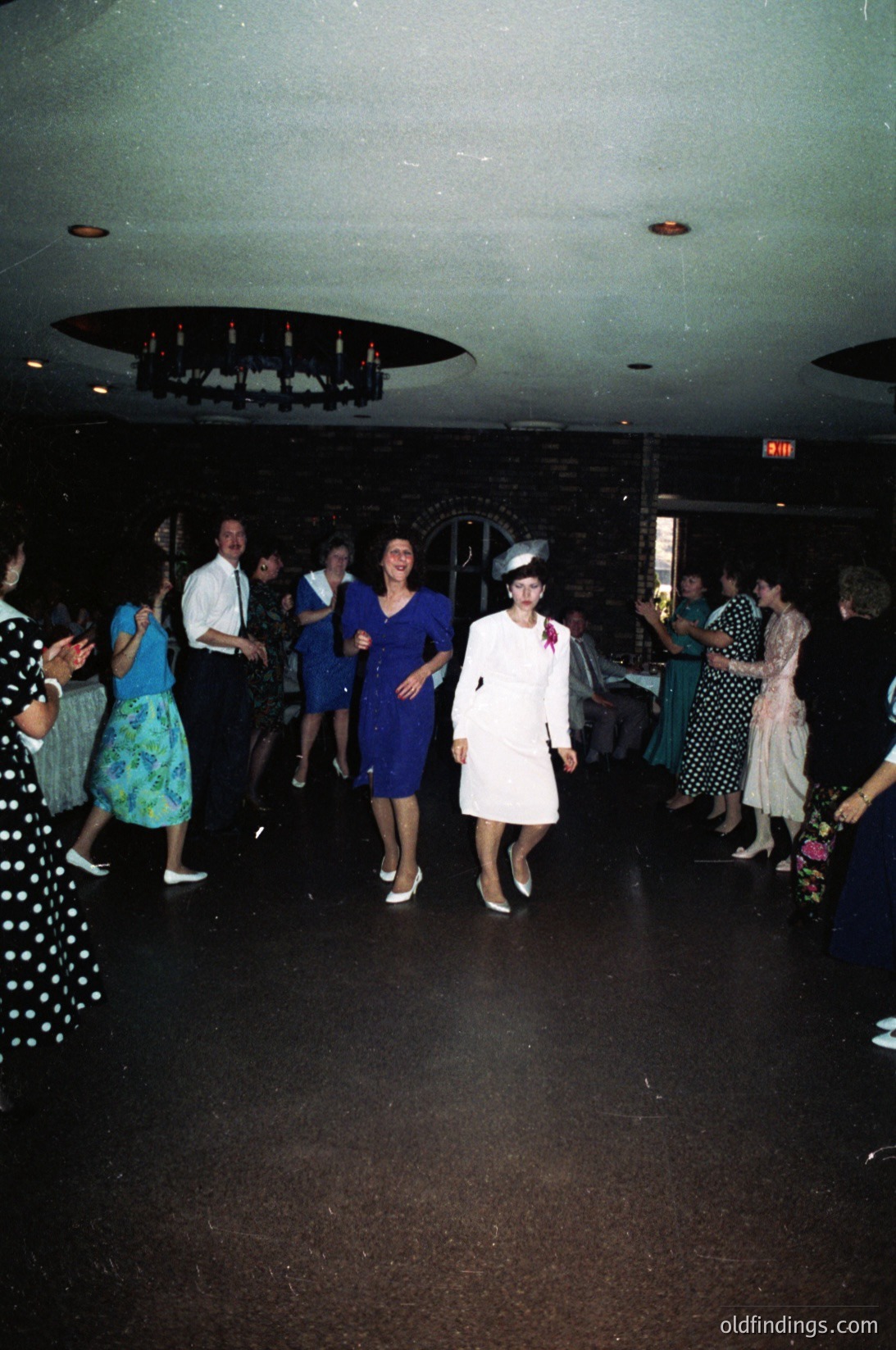 Vintage 1970s indoor dance party with retro attire—men in dress shirts, women in bold floral prints and fitted dresses. Central figure in white dress with feathered hat and pink sash leads the group. Circular chandelier and dark wood paneling suggest a mid-century venue. Candid, lively atmosphere captures era’s social energy.