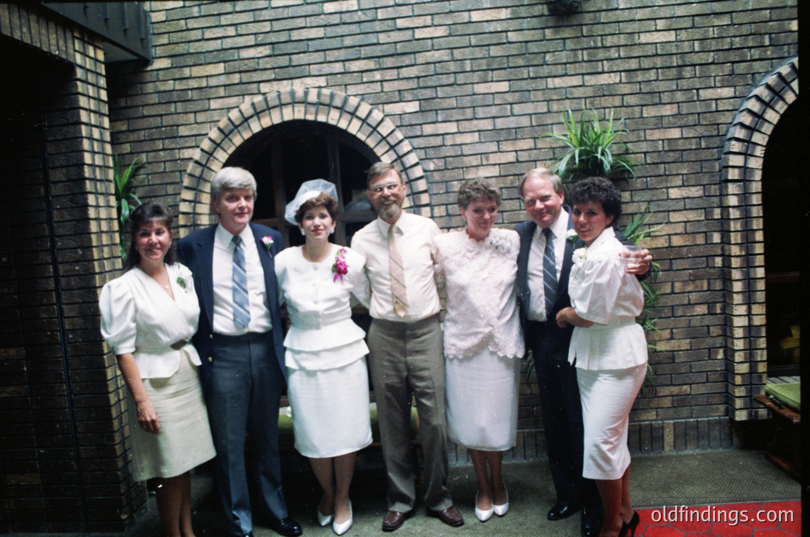 Six individuals pose in formal attire outside a brick building with an arched doorway, likely a 1970s wedding. Four women in white dresses with floral accents, two men in suits, and one in a nurse’s uniform. Decorative potted plant in foreground.