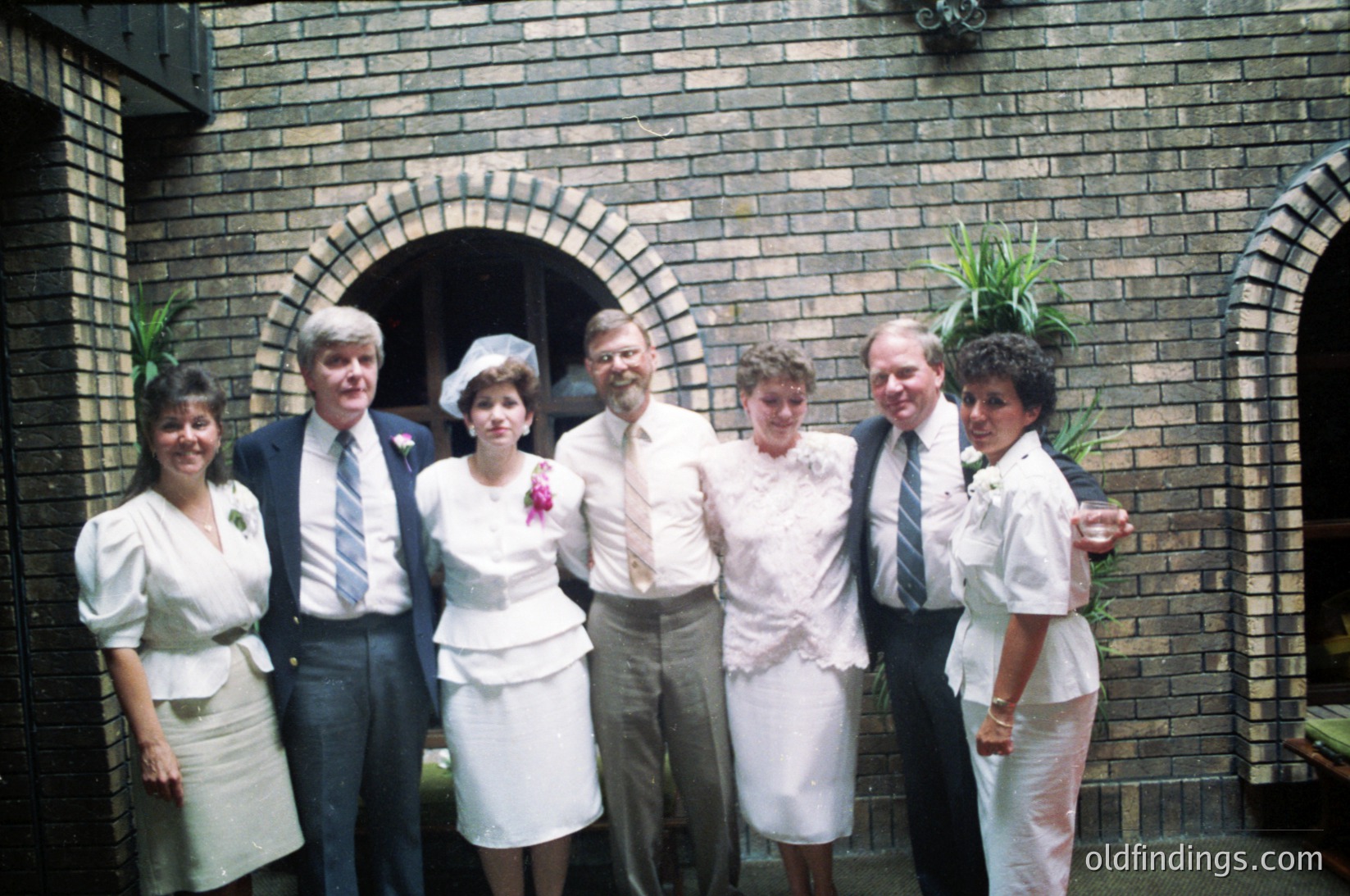 Six individuals pose outdoors in formal attire, likely a wedding or celebratory event, framed by a brick archway. The woman in the center wears a bridal gown with a bouquet; others in matching white dresses and suits. Floral headpieces and boutonnieres add detail. Mid-20th century (1960s–1970s) styling and brickwork suggest a European or North American setting.