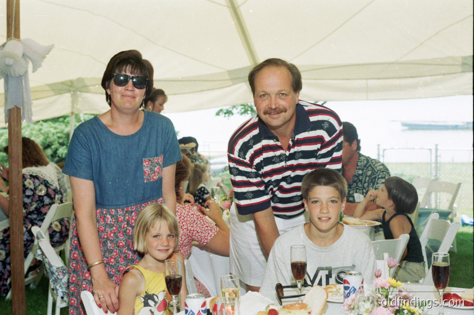 Family gathering at an outdoor event under a white canopy tent, likely a wedding or reception. Adults and children pose in 1980s-1990s attire: woman in floral skirt/dress, man in striped polo, boy in white T-shirt. Table setting includes wine glasses, floral centerpieces, and a "MIS" branded cup. Casual, festive atmosphere with blurred background guests.