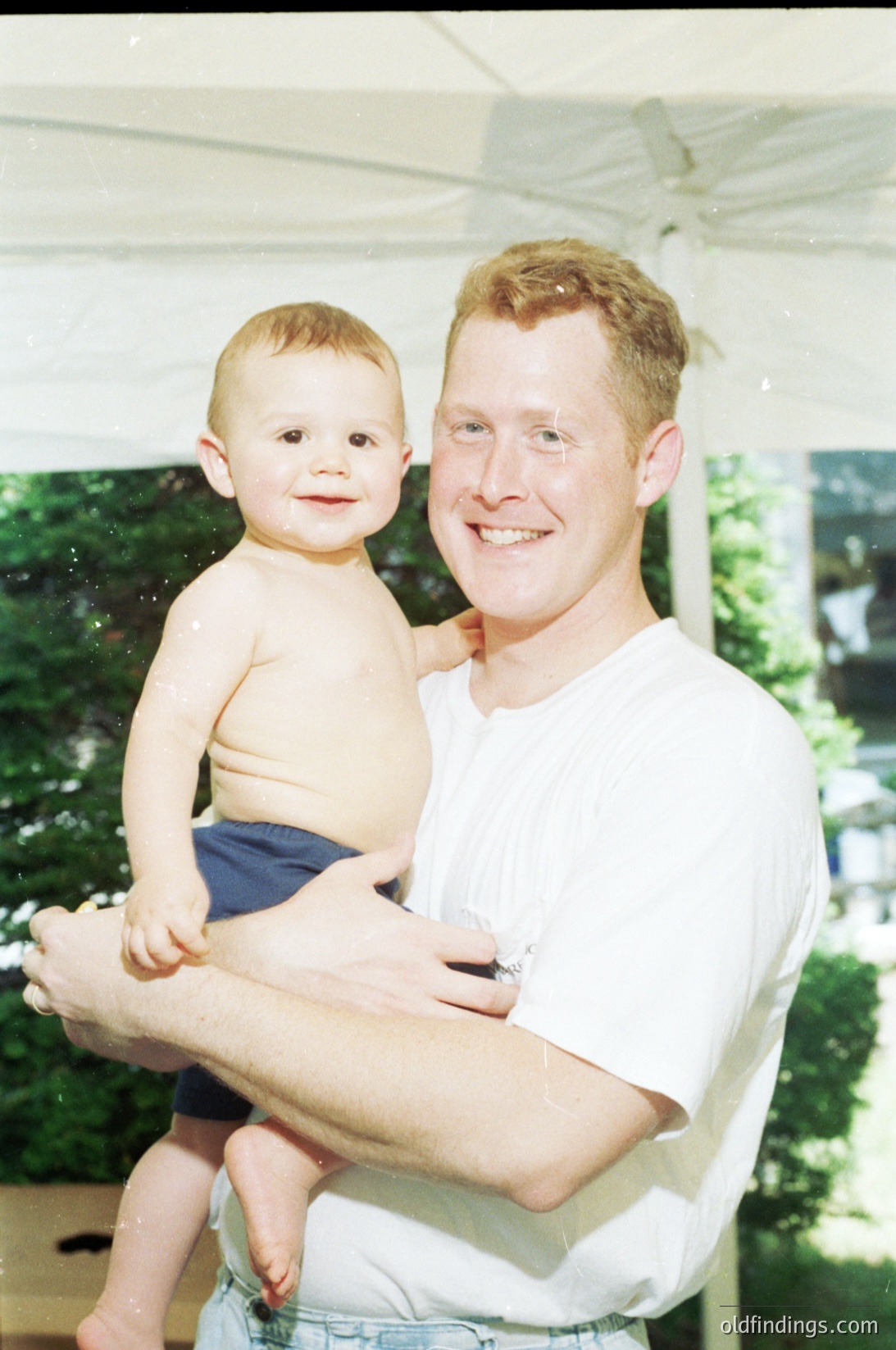 A man in a white shirt and denim shorts holds a toddler outdoors under a canopy. The child, shirtless in dark shorts, smiles directly at the camera. Greenery and a blurred background suggest a garden or patio setting, likely mid-2000s.