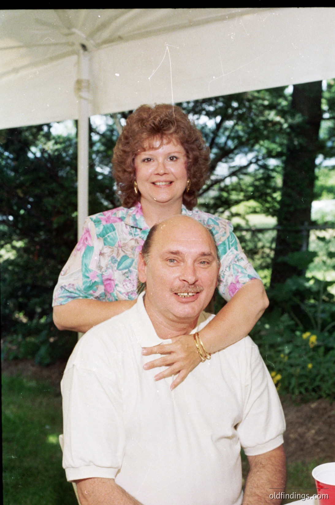 A woman in a floral-patterned blouse stands behind a smiling man in a white polo shirt under a tent, likely at an outdoor event. The vintage digital quality suggests the photo was taken in the **1990s–2000s**. The man’s gold watch and casual pose indicate a relaxed, informal gathering.