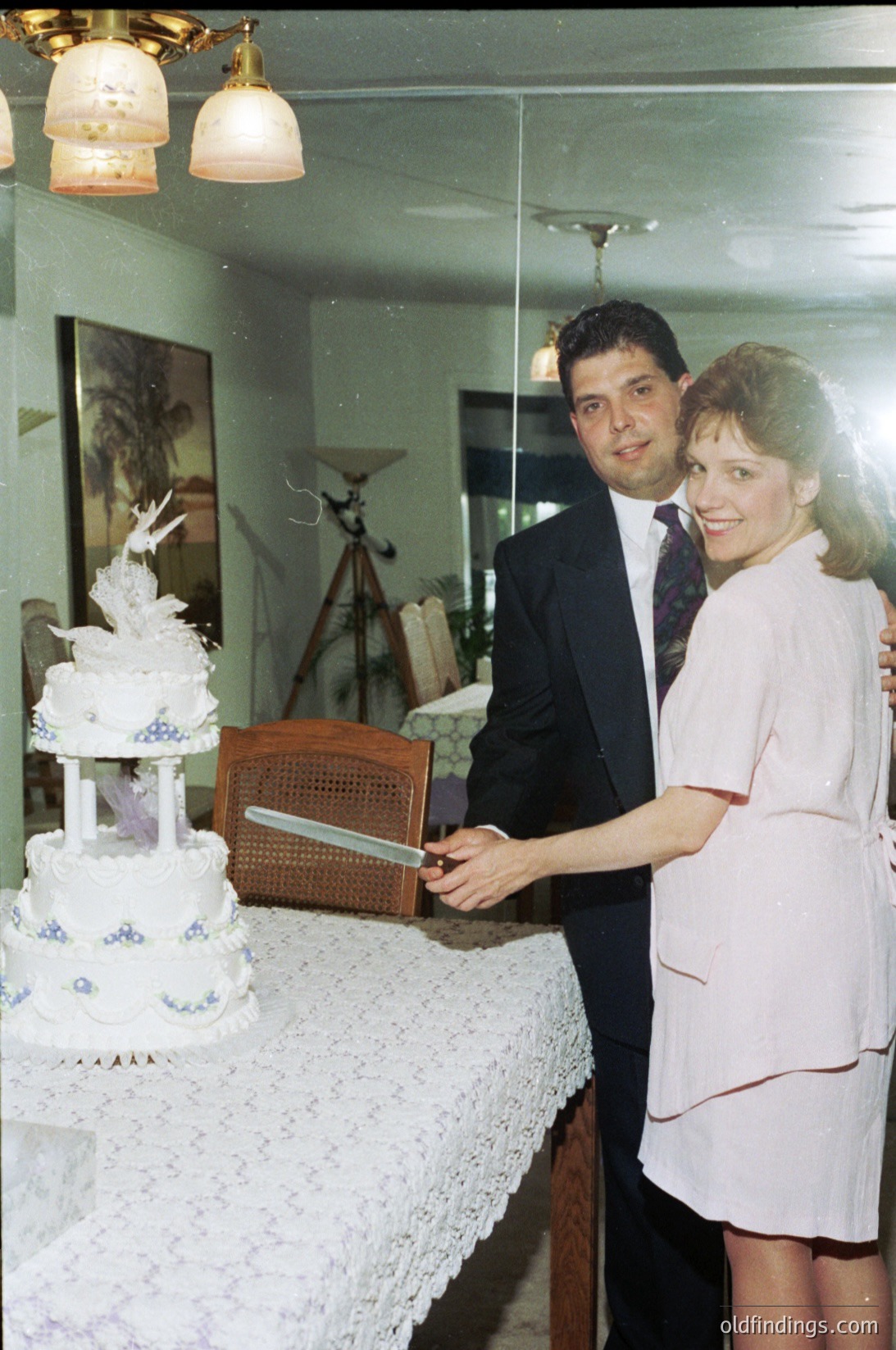 A 1970s-style wedding cake cutting scene in a vintage home. The multi-tiered cake features blue floral designs and a bird motif. The man in a dark suit and woman in a white gown pose beside a wooden rocking chair and lace tablecloth. Classic interior with ornate ceiling fixture and framed artwork.