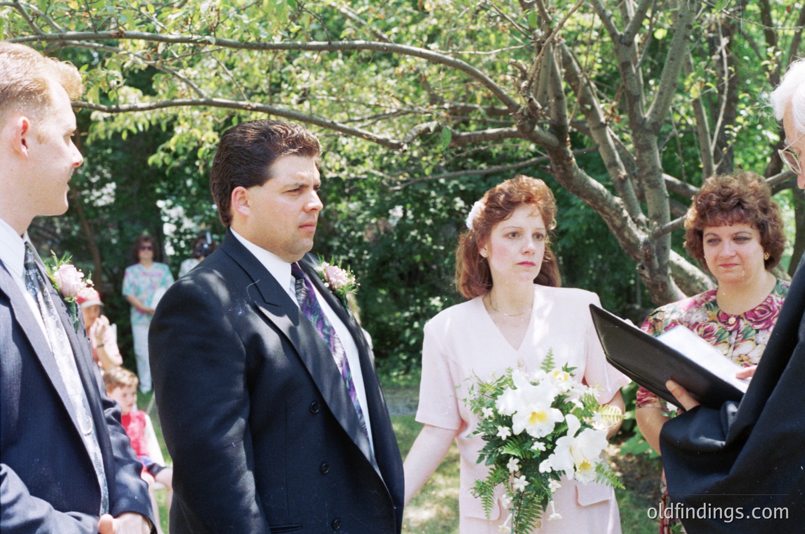 1990s outdoor wedding ceremony with bride in vintage-style dress holding white flowers. Groom in dark suit, officiant in floral blouse reading from a book. Lush greenery and natural light create a serene backdrop.