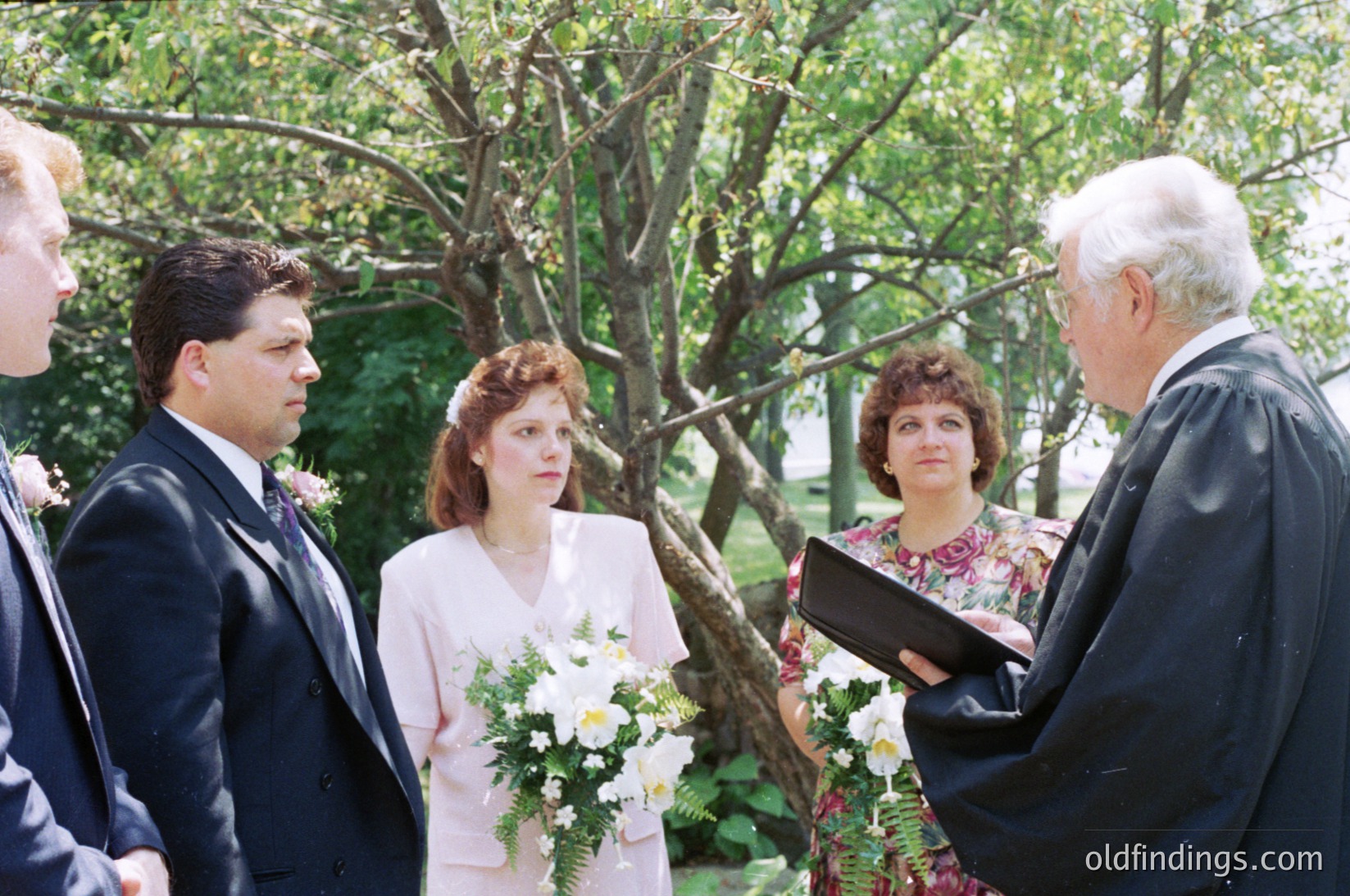 Outdoor wedding ceremony with bride in vintage 1990s attire holding white bouquet. Officiant in black robe conducts ceremony under leafy trees. Formal suits and floral dresses suggest mid-to-late 1990s U.S. wedding traditions.