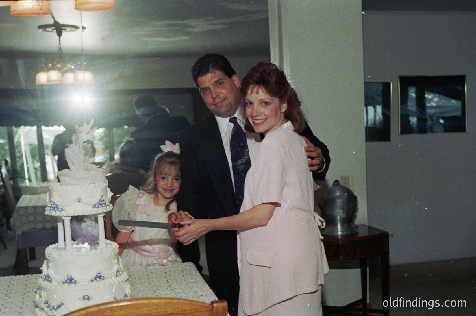 Family wedding celebration with three generations: bride (white lace gown), groom (dark suit), and young girl (dress with ribbon) cutting a multi-tiered cake adorned with blue floral accents. Indoor venue features vintage decor, including a chandelier and retro appliances. Likely 1980s–1990s Western setting.