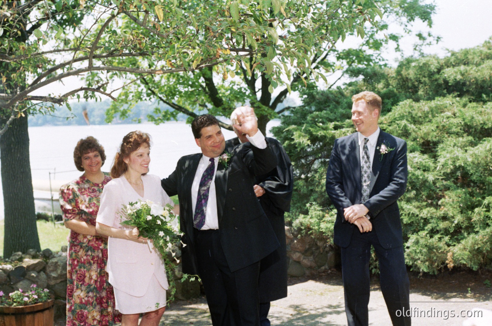 Outdoor wedding ceremony by a serene lake, featuring four individuals in 1990s attire. The bride in a light floral dress holds a bouquet, flanked by a woman in floral-patterned dress and a man in a dark suit raising his hand. The groom and best man in dark suits with boutonnieres stand beside them. Lush greenery and water backdrop suggest a lakeside venue.