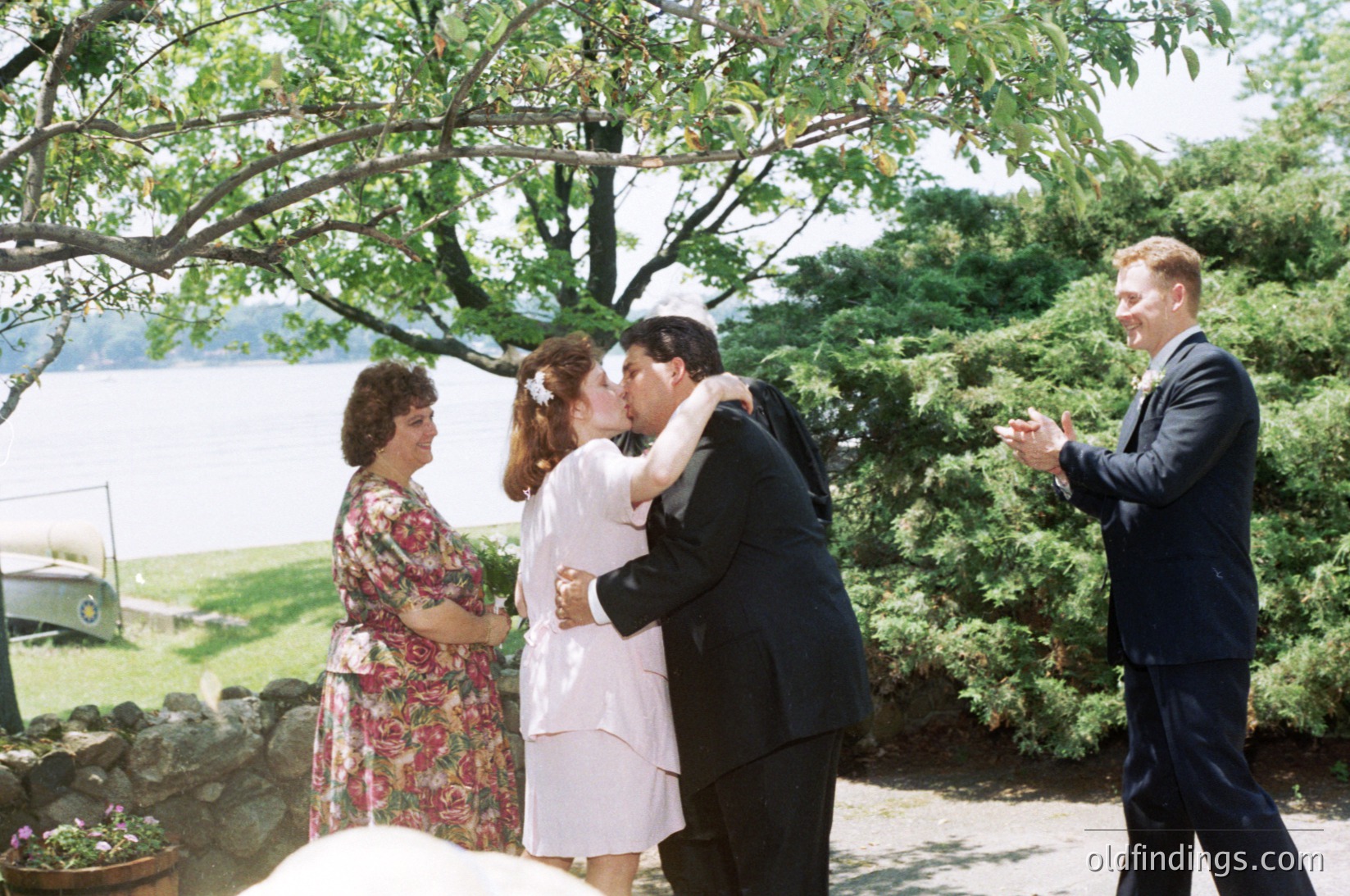 A 1970s lakeside wedding scene: bride and groom share an emotional embrace under a leafy tree, surrounded by floral dresses and dark suits. Witnesses pose nearby—one in floral print, another in a classic suit. Vintage car parked in background suggests a rural or suburban lakeside venue.