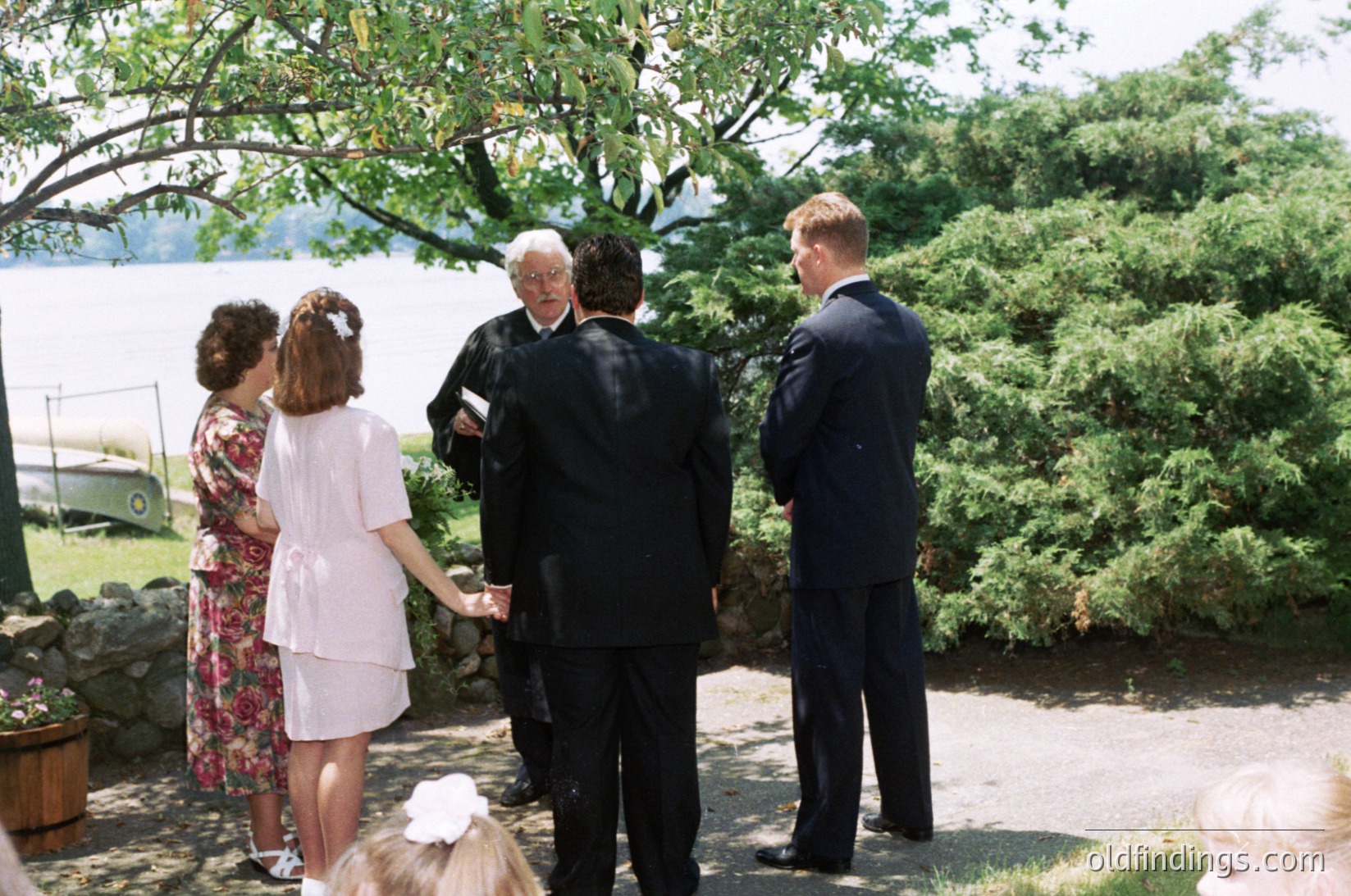 Five adults in formal attire—men in suits, women in dresses—gather outdoors near a serene lake, likely for a wedding or ceremony. Lush greenery, stonework, and a vintage vehicle in the background suggest a mid-20th-century estate setting ( ).
