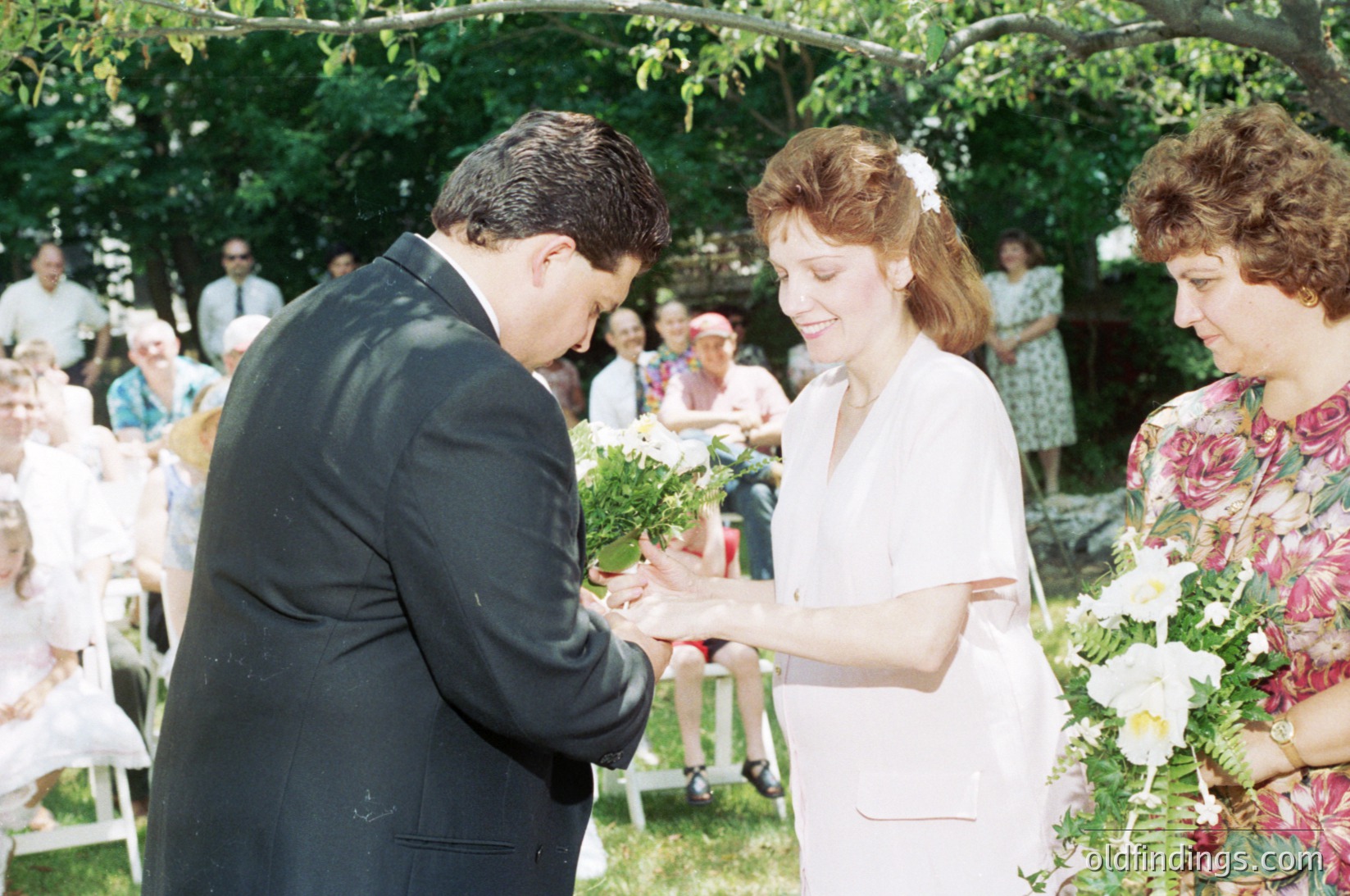 1970s outdoor wedding ceremony with bride and groom exchanging bouquets. The groom in a dark suit and the bride in a white gown with floral headpiece. Guests seated at white tables under trees. Floral arrangements and vintage attire suggest a mid-century European wedding.