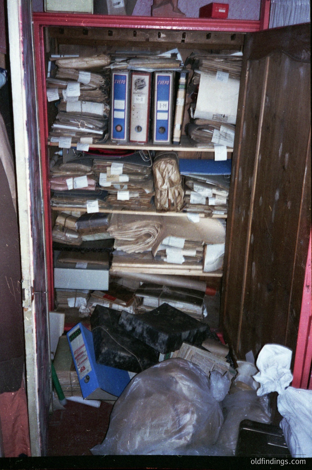 Overcrowded metal filing cabinet filled with aged binders, loose documents, and miscellaneous paper stacks. Evidence of decades-old archival storage, likely institutional or office-based. Possible Soviet-era or Eastern Bloc setting (1960s–1990s).