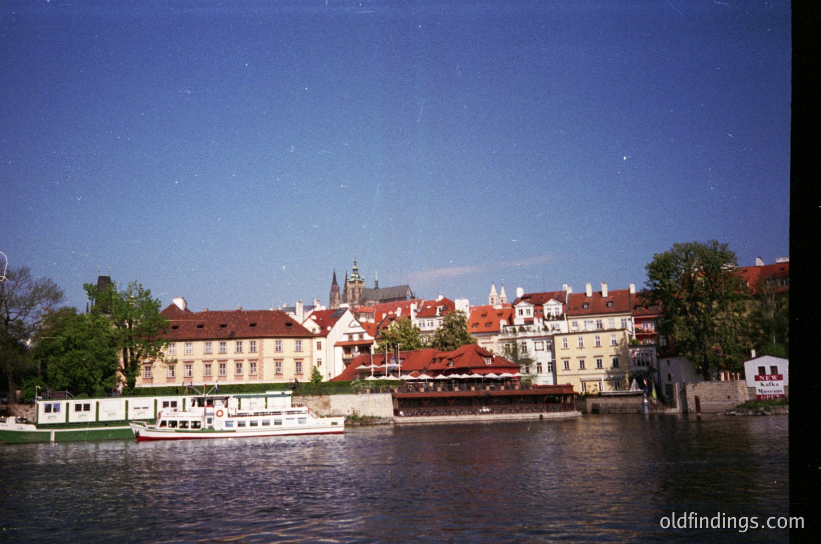 Historic European riverside scene with classic red-tiled rooftops and a prominent church spire. Mid-20th century architecture along a calm waterway, likely Prague’s Vltava River. Vintage boat docked near a riverside café or restaurant. é