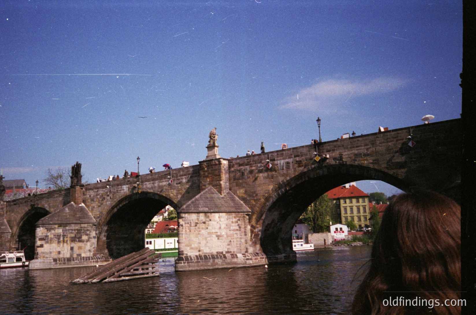 Vintage photograph of the historic **Charles Bridge (Karlův most)** in Prague, Czech Republic, featuring Gothic stone arches and statues. The bridge spans the Vltava River, with boats and pedestrians visible. Distinctive medieval architecture and vintage filter enhance nostalgic appeal.