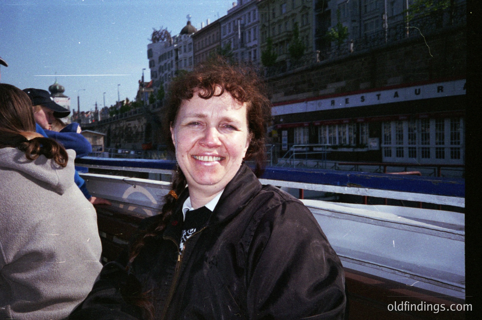 Woman in a dark jacket smiles outdoors near a riverboat labeled "Hestur." Historic European architecture with ornate facades and domed roofs in background. Likely 1990s–2000s, Scandinavian or Baltic region.