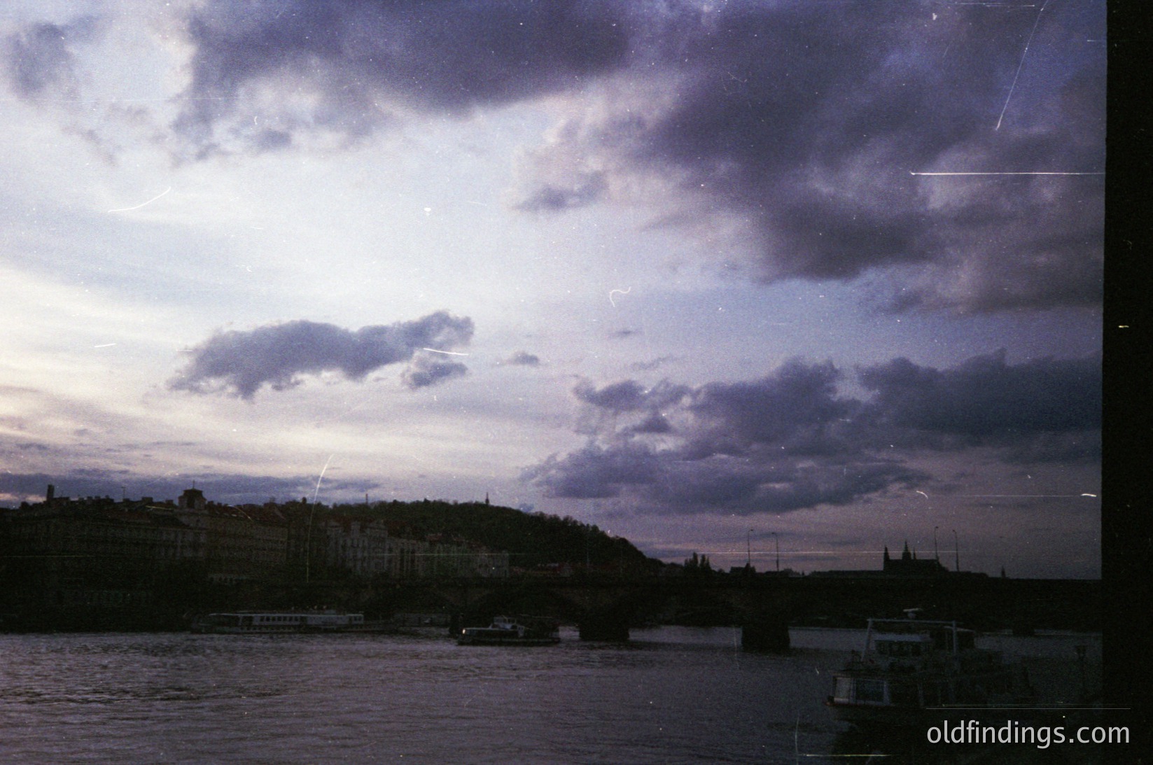 Vintage urban riverscape at dusk, featuring a bridge spanning a calm waterway. Dense, low-rise buildings line the hillside, suggesting a European cityscape. A lone boat floats near the shore, while another crosses the bridge. Dramatic storm clouds dominate the sky, casting moody lighting. Likely 1970s–1980s due to film grain and color tone.