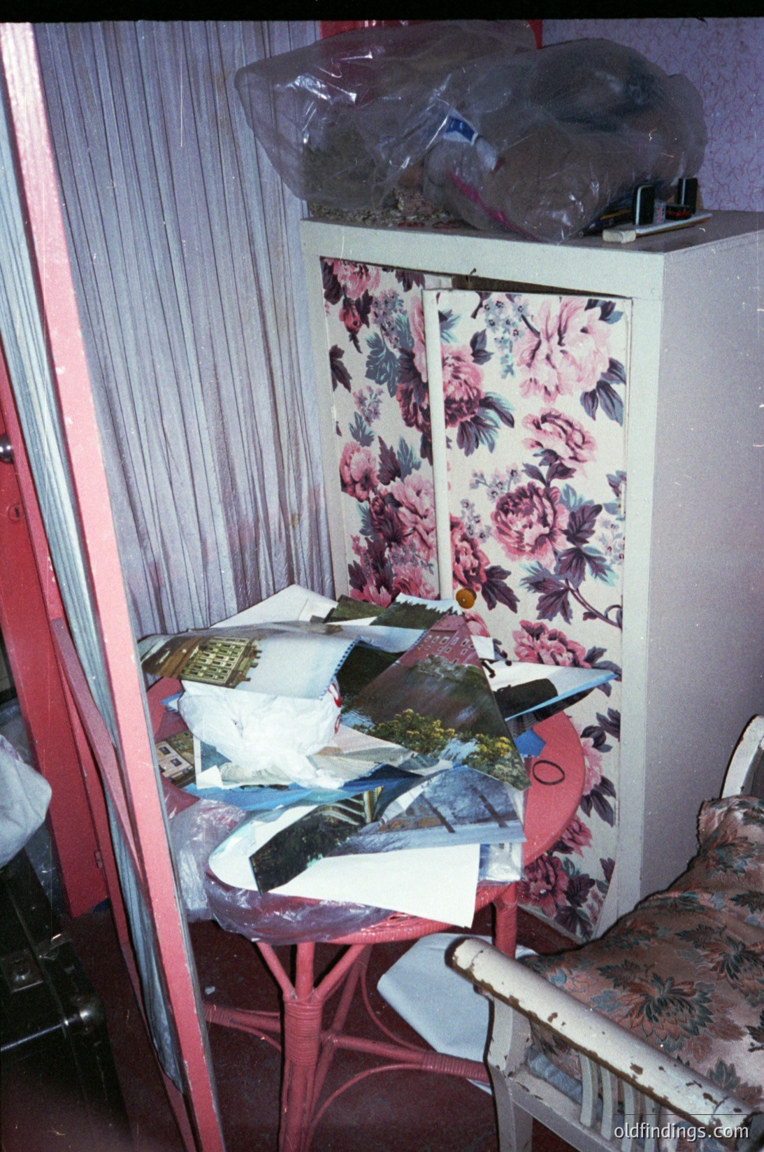 Vintage floral-patterned cabinet with floral wallpaper backdrop, displaying folded garments and textiles. Mid-century design with red metal frame. Possible 1960s–1980s domestic setting.
