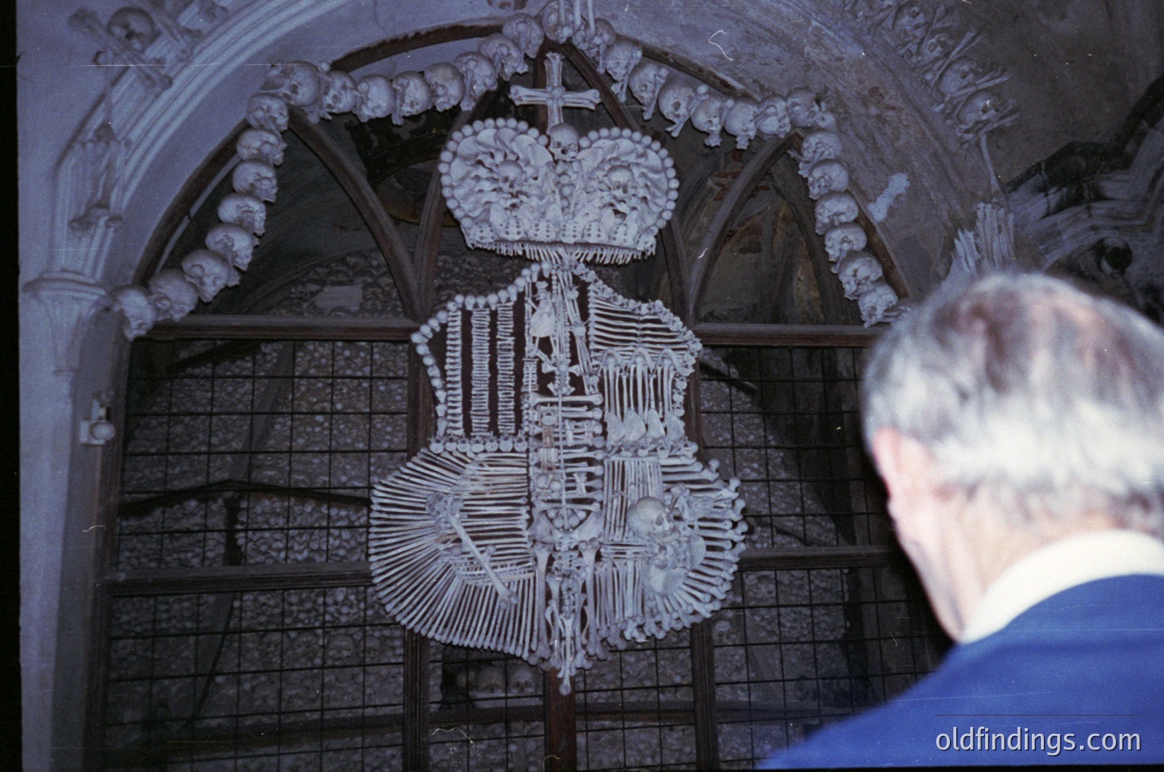 Bone altar piece featuring a crucifix and coat of arms, framed by Gothic-style arches. Likely a 16th–18th century European ossuary or chapel interior.