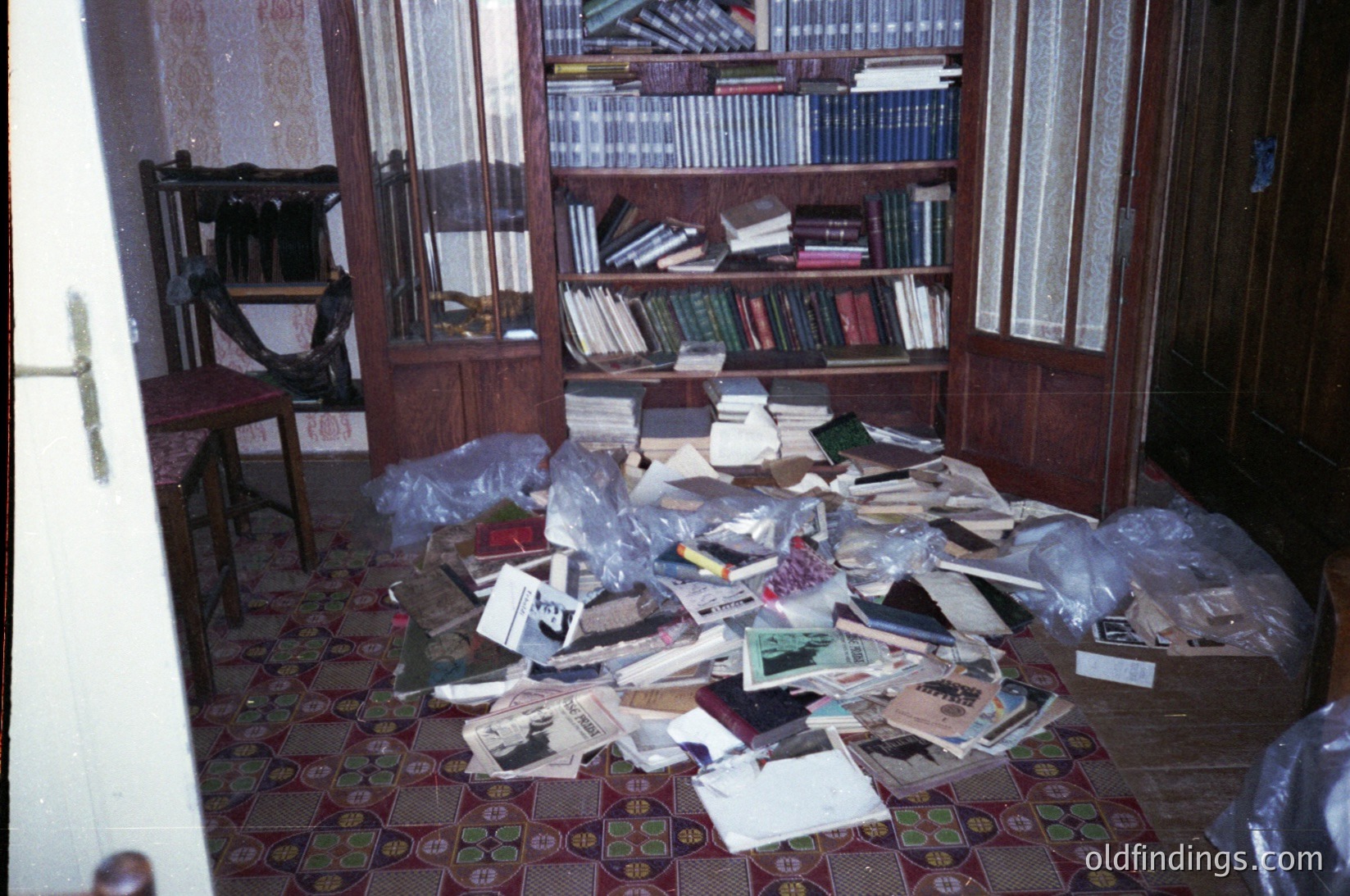 Vintage room in disarray, featuring wooden paneling and a tiled floor with floral patterns. Shelves overflow with books, mixed with scattered papers, envelopes, and plastic bags. Likely a mid-20th century domestic setting, possibly Eastern European.