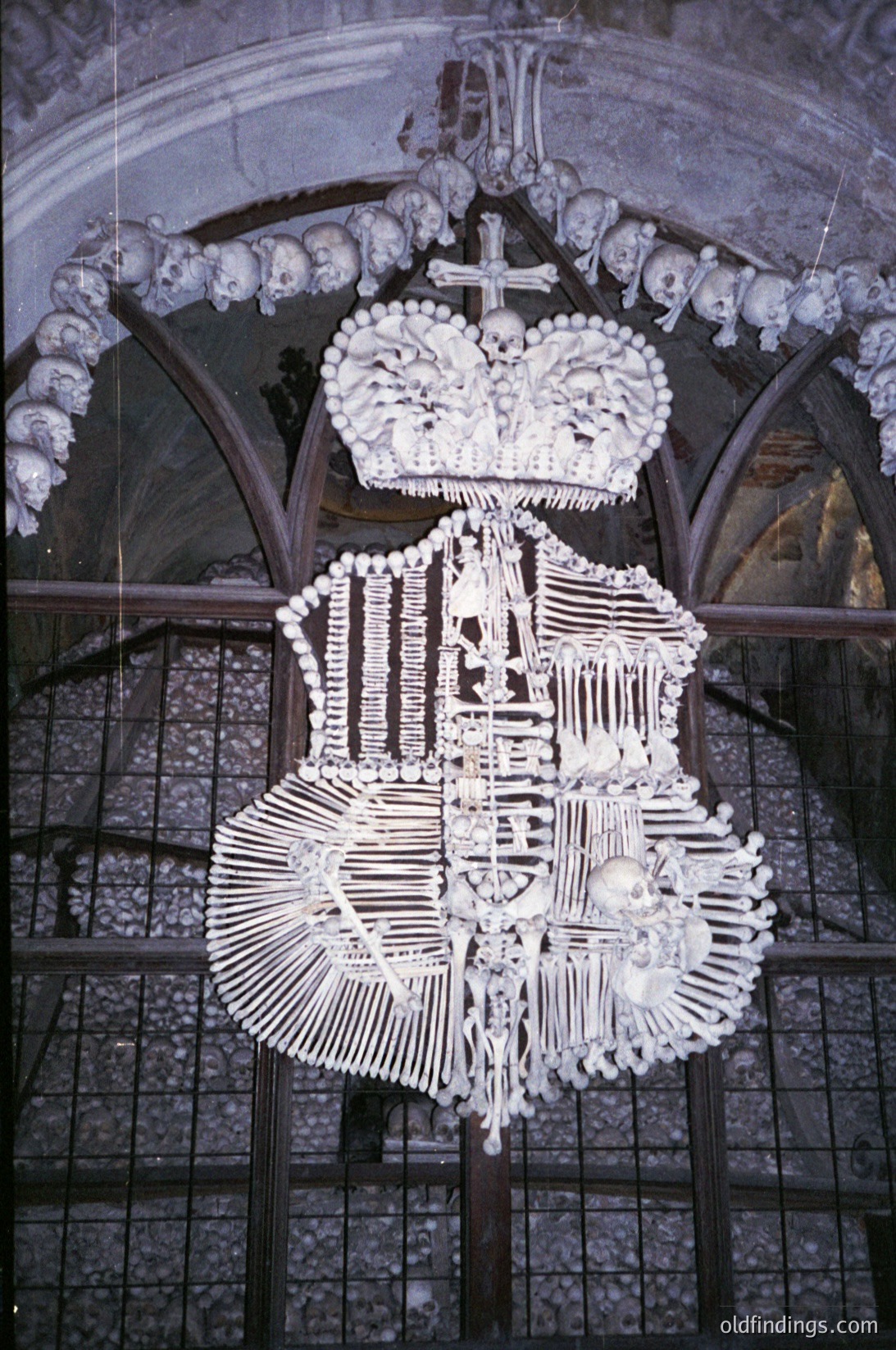 Bone chandelier crafted from human remains, featuring a crown, cross, and ornate religious motifs. Hanging in a Gothic-style chapel with skulls embedded in the arch. Likely from a 19th-century European ossuary, possibly (á Hora). Unique historical artifact blending macabre artistry with religious symbolism.