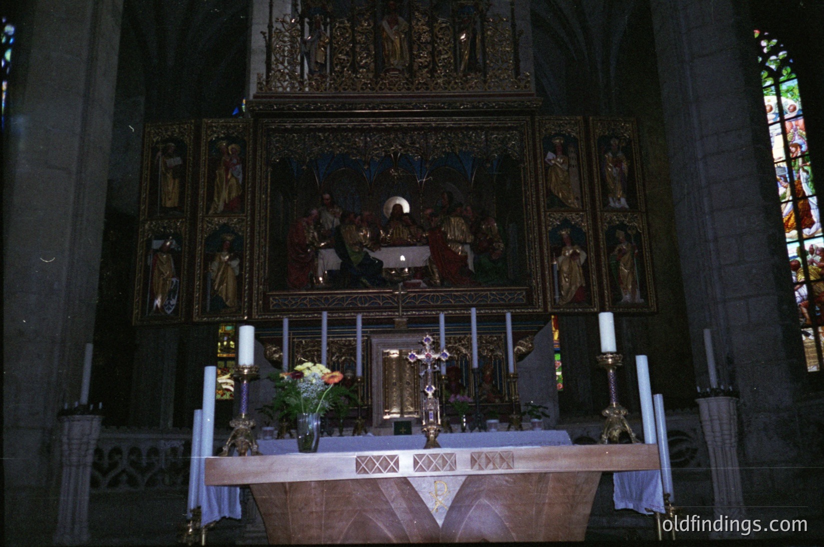 Gothic-style altar with intricate wooden carvings, flanked by candle holders and floral arrangements. Central mural depicts religious figures in a liturgical scene. Stained-glass windows with geometric patterns in background. Likely European cathedral, 19th–early 20th century.