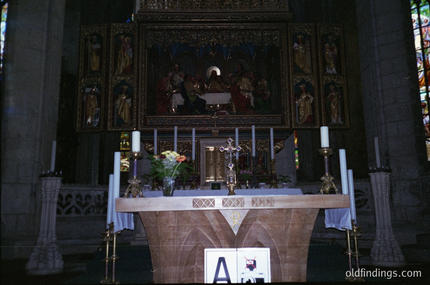 Gothic-style altar with ornate wooden carvings and central crucifix, flanked by candle holders and floral arrangements. Stained-glass windows and religious iconography depict biblical scenes. Likely European cathedral, 19th–early 20th century.