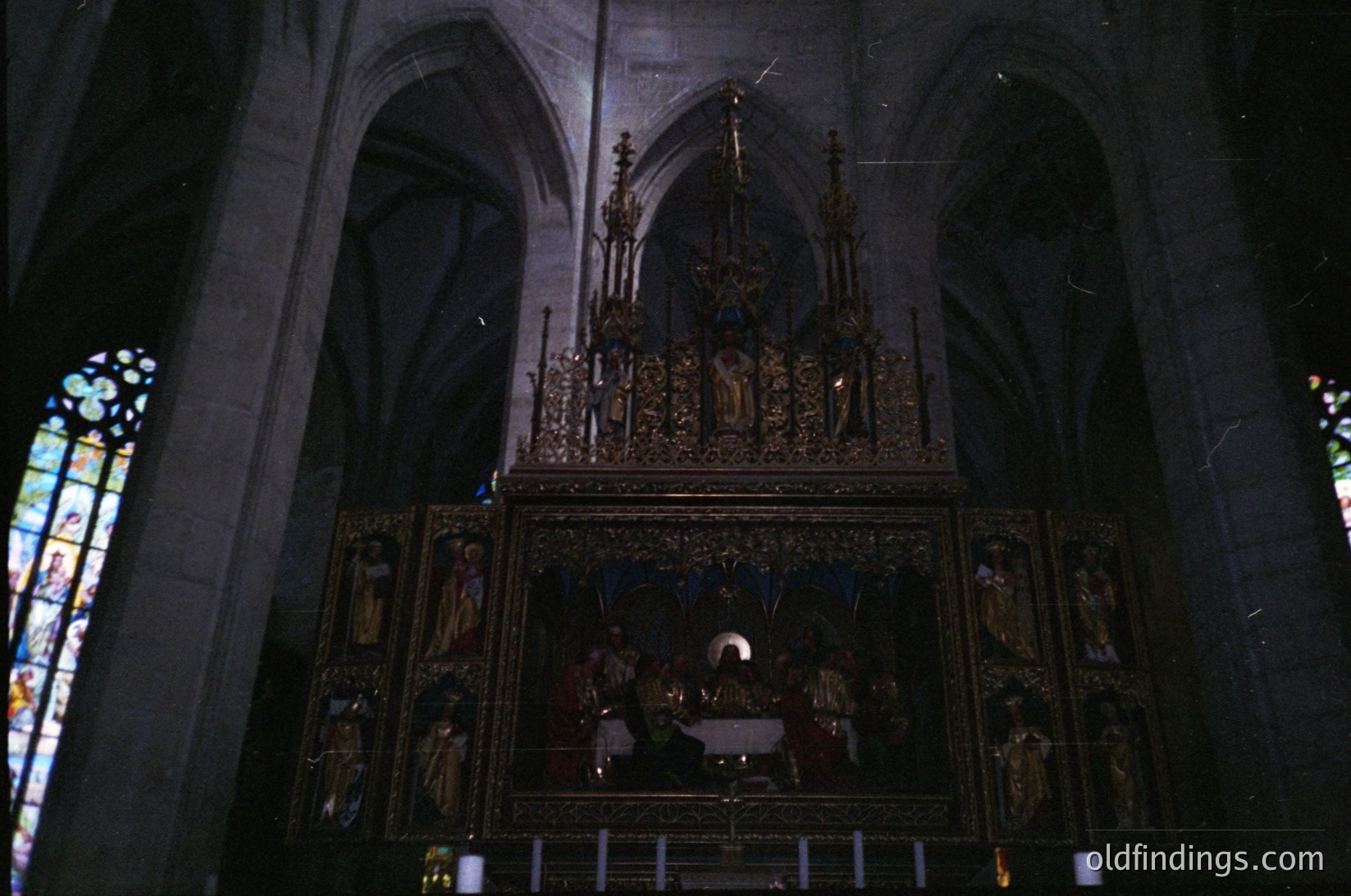 Gothic-style altar with ornate wooden carvings depicting religious scenes, flanked by stained-glass windows. Intricate detailing includes crucifixion motifs and robed figures. Likely European, 12th–16th century.