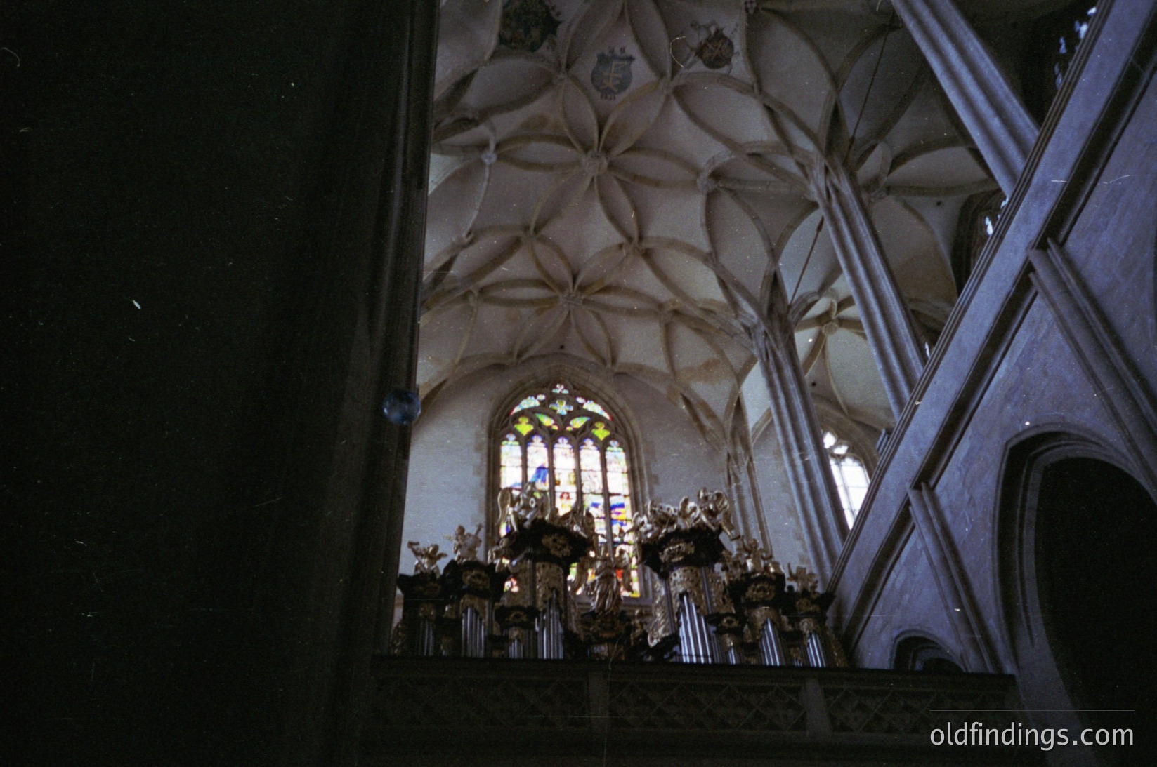 Gothic-style cathedral interior with ribbed vaulted ceiling and stained-glass window. Ornate altar featuring carved wood and religious iconography. Light filtering through stained glass creates colorful patterns on stone walls. Likely European, 13th–16th century.