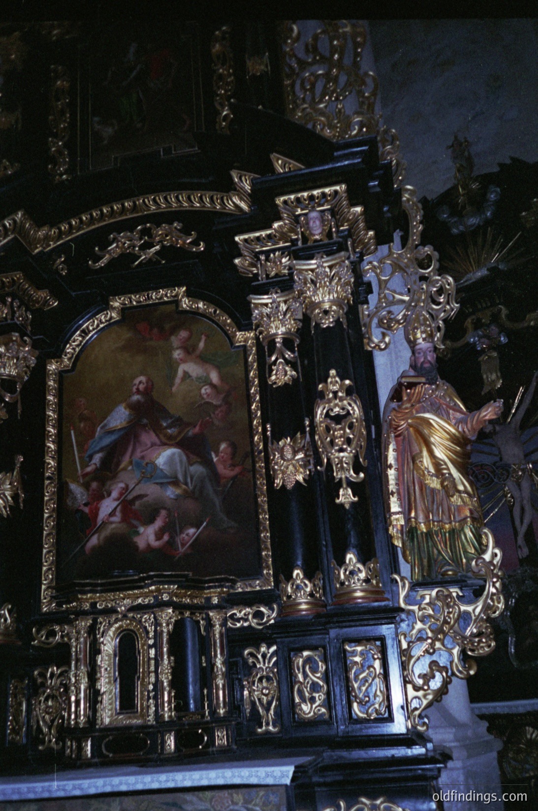 Baroque-style altar featuring gilded woodwork, intricate carvings, and a central religious painting depicting saints. Ornate columns, sculpted angels, and decorative motifs dominate the scene. Likely European, 17th–18th century.