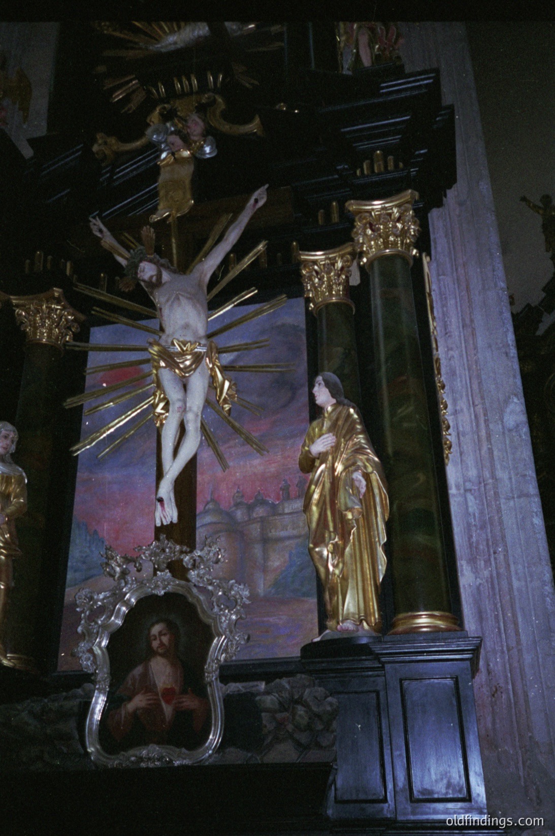 Baroque-style altar featuring a crucifixion scene with gilded accents. Central crucifix with radiating light rays, flanked by a golden statue of the Virgin Mary in mourning pose. Intricate floral and scrollwork details on the altar frame. Painted portrait inset below the crucifix. Likely European, 17th–18th century.