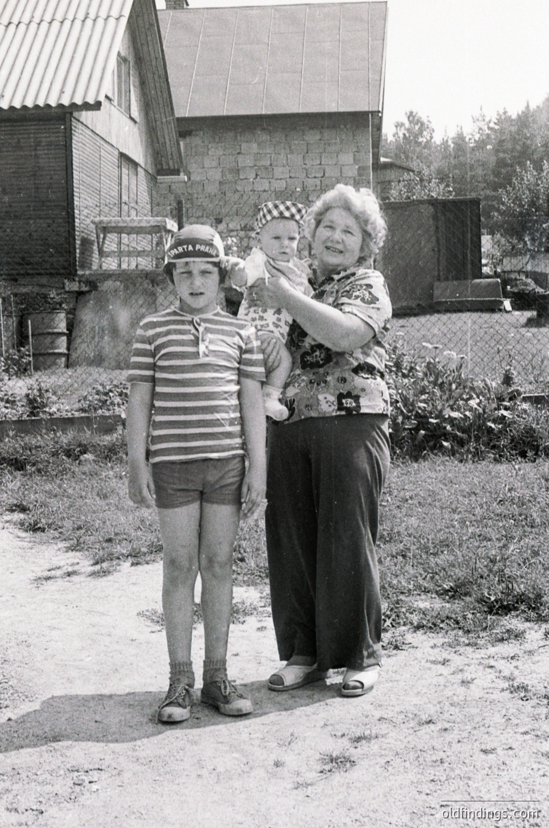 A black-and-white snapshot from the 1960s–70s featuring a grandmother, child, and baby outdoors in a rural setting. The woman wears a checkered headscarf and floral blouse, holding a baby in a striped outfit. The boy stands beside her in a striped polo shirt and shorts. Wooden house with a corrugated roof and rustic fence in background.