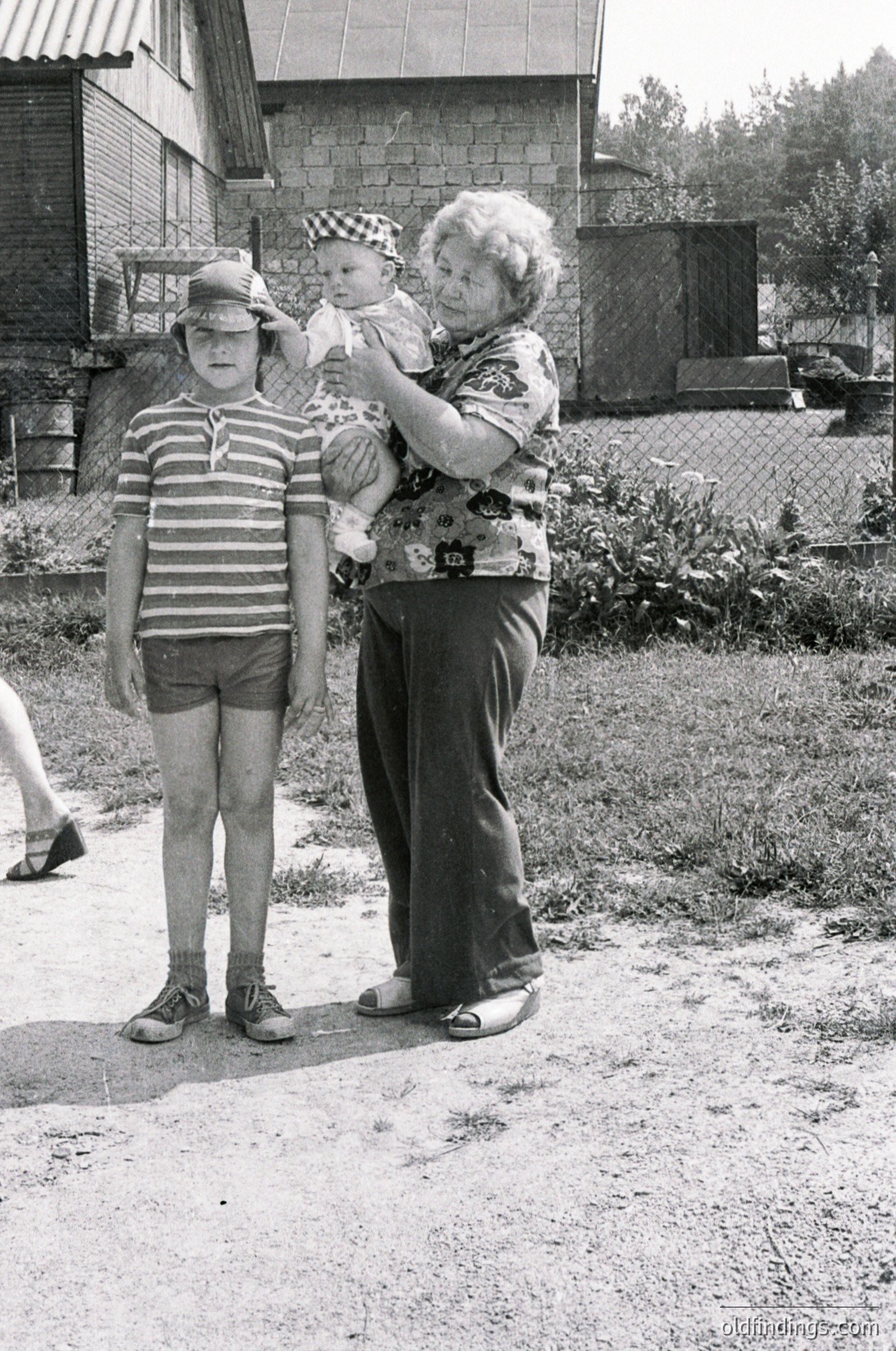 A mid-20th-century black-and-white snapshot captures a grandmother, child, and toddler in a residential backyard. The grandmother, wearing a floral blouse and dark skirt, holds a baby in a checkered hat. A young boy in a striped shirt and shorts stands beside her. The setting features a modest brick house, overgrown greenery, and a chain-link fence. [Vintage family portrait ]