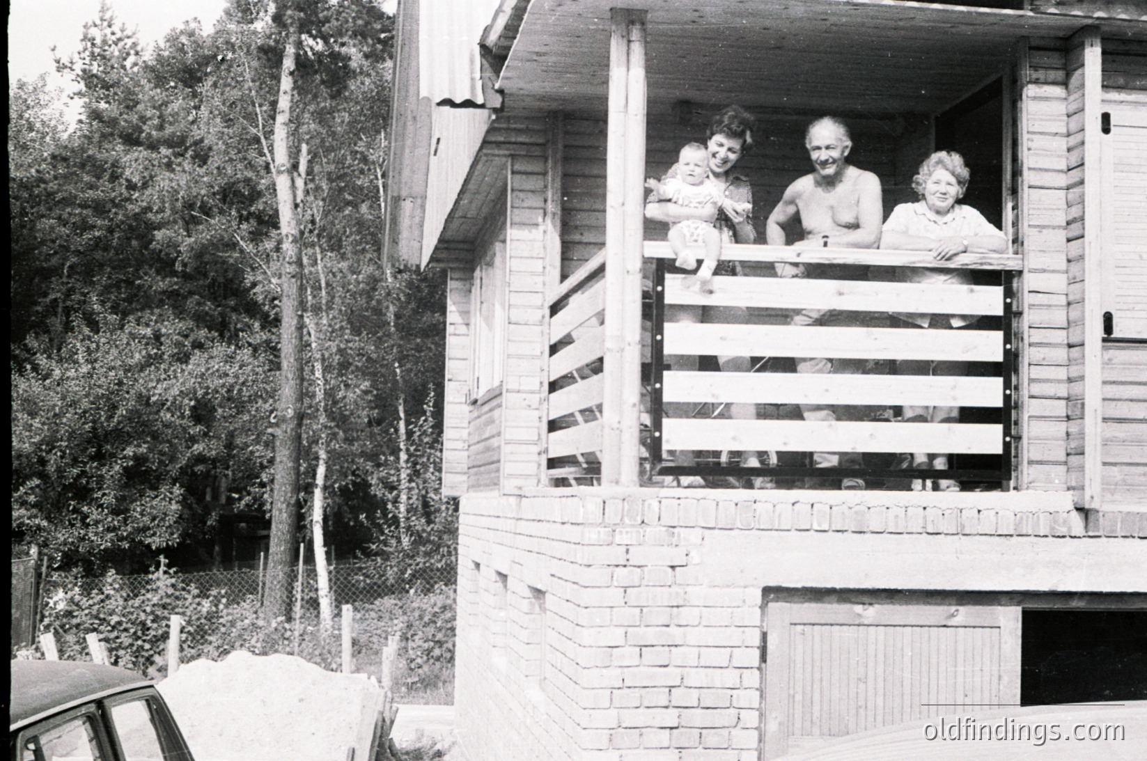 Three adults pose on a wooden balcony of a brick house, mid-20th century. Lush greenery and a parked car suggest a suburban or rural setting.