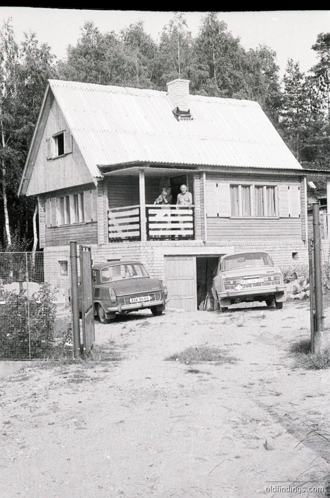A 1960s-era wooden cabin with gabled roof and balcony, surrounded by dense forest. Two vintage cars (likely Lada or similar) parked under a covered garage entrance. A child and adult visible on balcony, suggesting a rural or suburban setting. Black-and-white photo captures mid-century architectural style and lifestyle.
