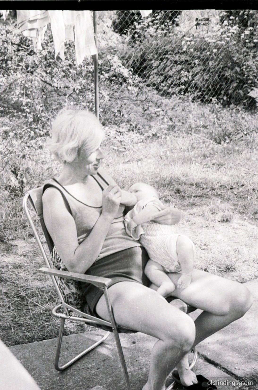 A woman in a sleeveless swimsuit cradles a small white dog in a metal folding chair, mid-1960s outdoor setting. Lush greenery and a flagpole with a faded banner in the background suggest a garden or park. Classic mid-century fashion and pet portraiture.