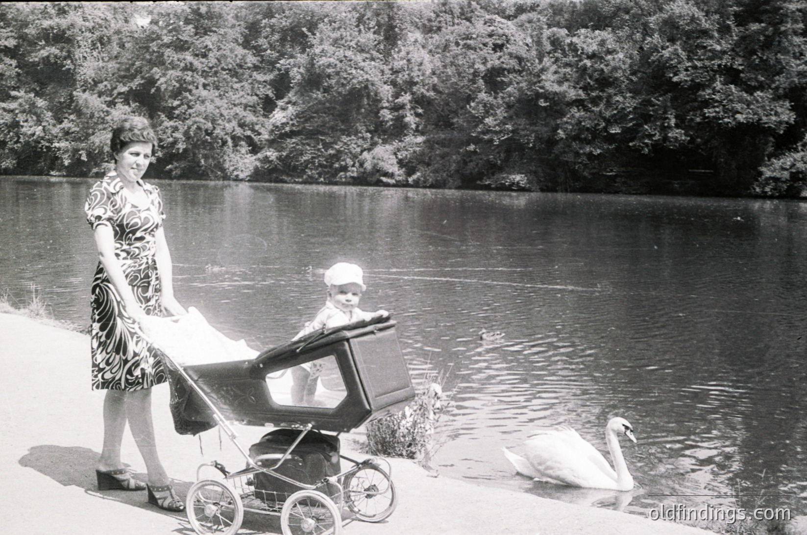 Mid-20th century black-and-white photo: woman in patterned dress pushes vintage pram near serene lake, swan glides in background. Lush forested shoreline suggests park or countryside setting.