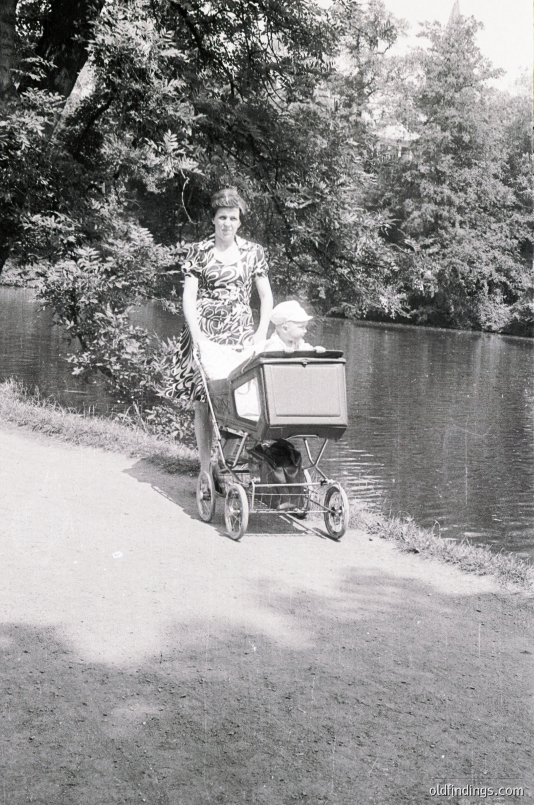 A woman in a patterned dress pushes a vintage baby carriage along a riverside path, mid-20th century. Lush greenery and water reflect the serene outdoor setting.